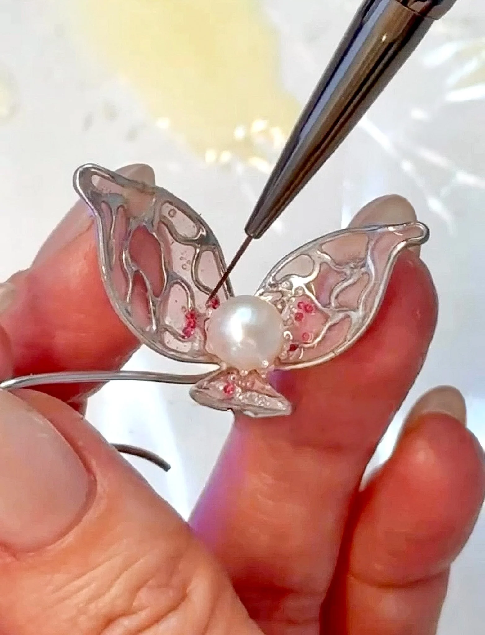 Close-up of a person's hand holding a decorative butterfly-shaped object made of transparent material with red accents, and a pearl-like bead at the center, with metallic tools in the background.