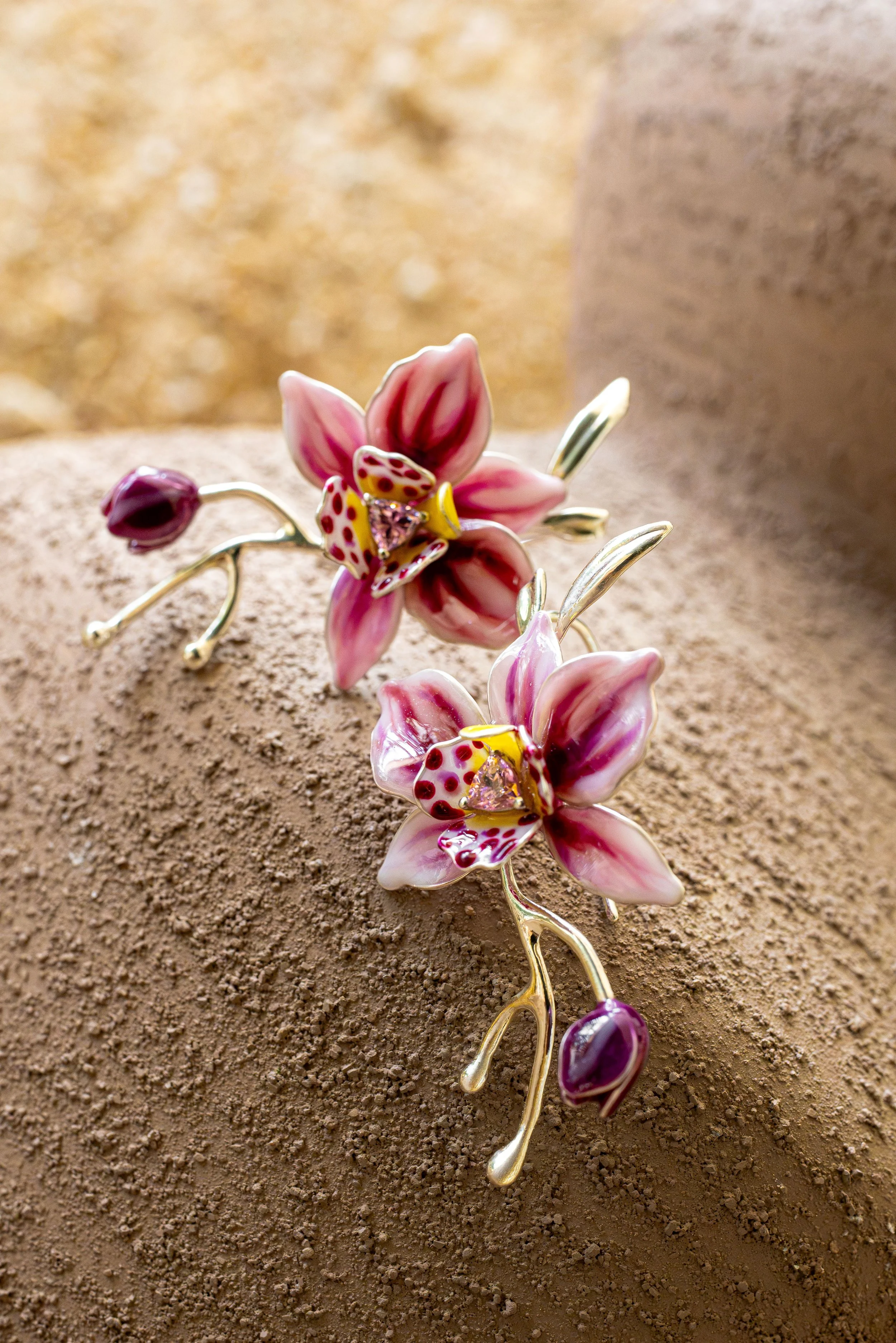A decorative pink and gold orchid-shaped jewelry piece with gem and enamel details, resting on a textured beige surface with a blurred gold and beige background.