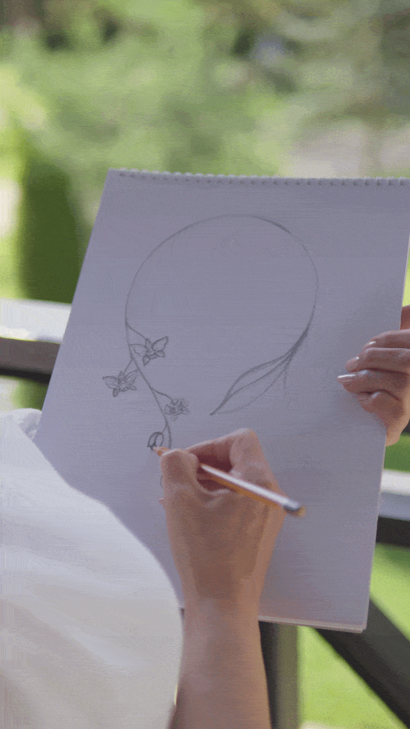 Person drawing a balloon with butterfly and flower details on a piece of paper, held up near window.