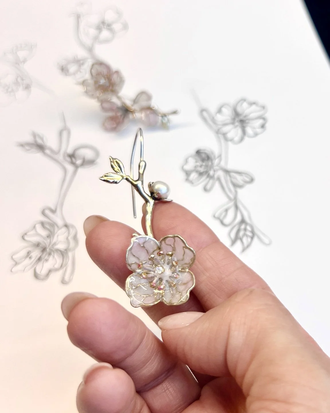 Close-up of a hand holding a flower-shaped gold jewelry piece with a pearl, with a background of shadow outlines of similar floral jewelry.