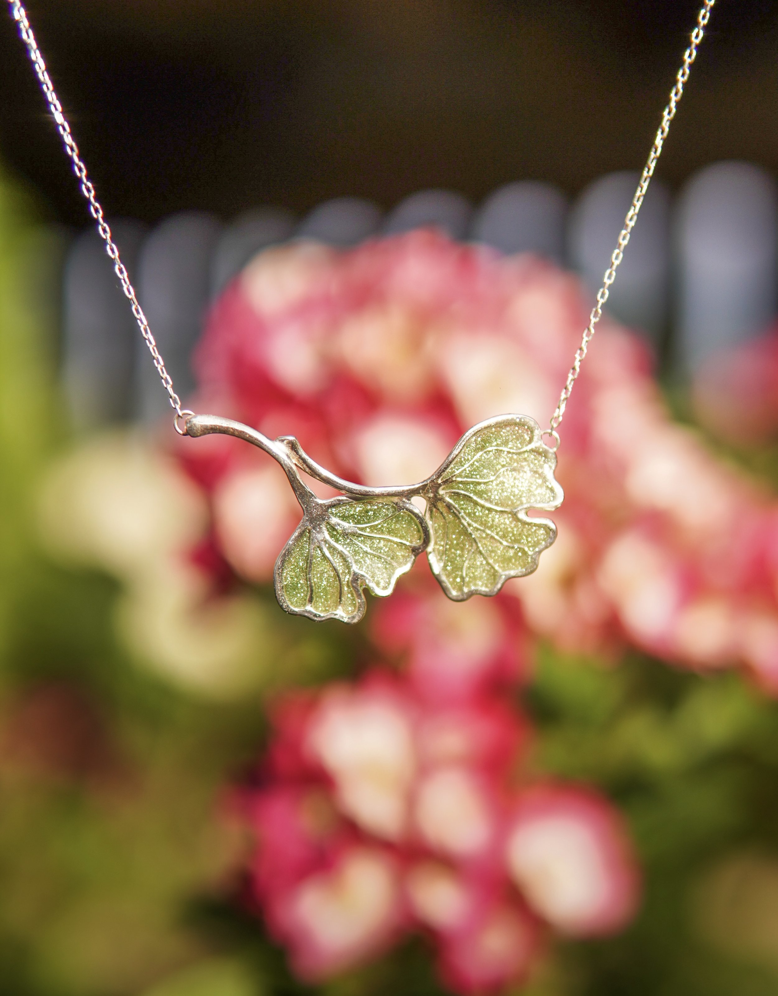 A close-up of a necklace with a butterfly-shaped pendant, hanging in front of a blurred background of pink and white flowers.
