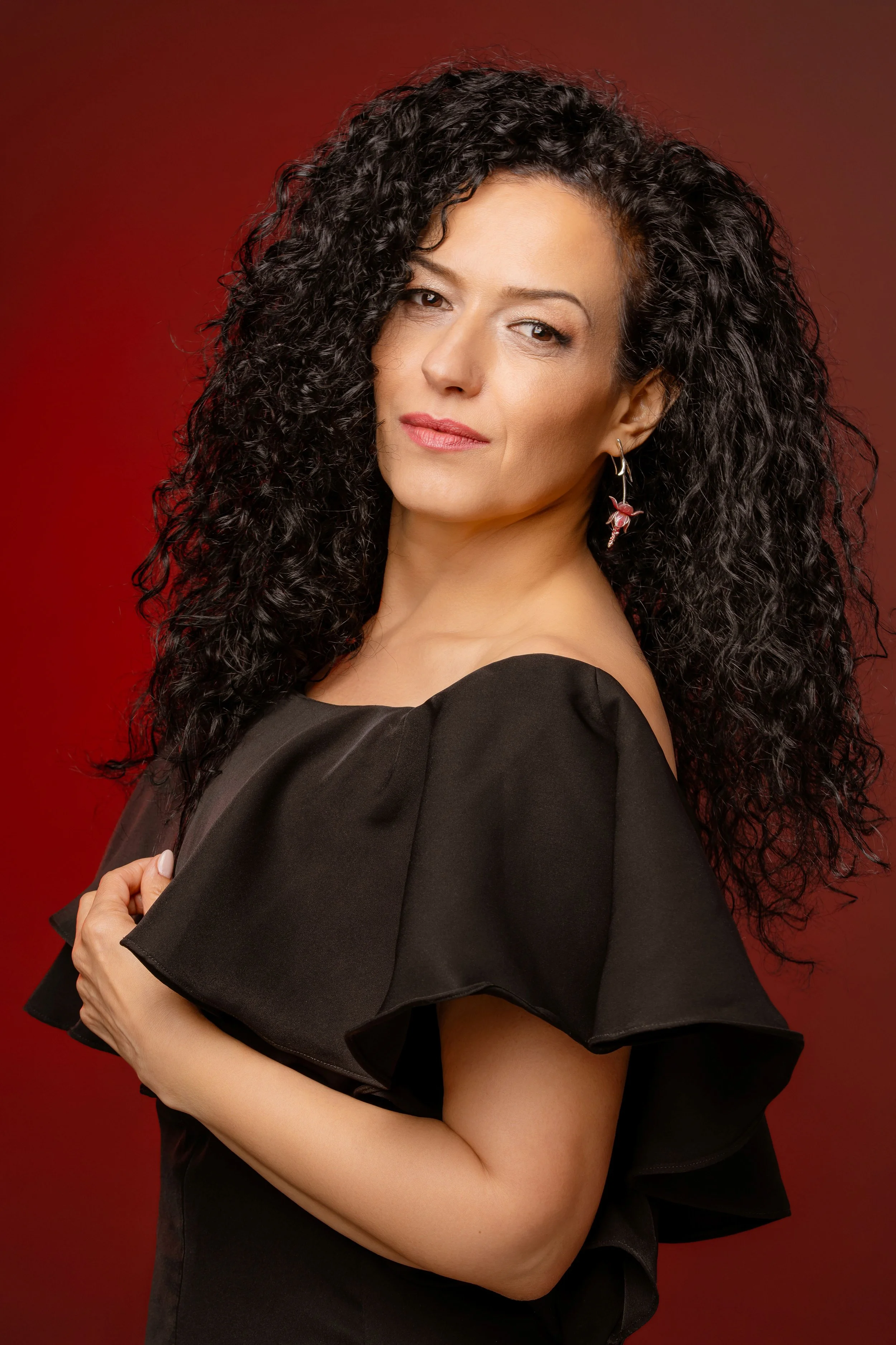 A woman with curly black hair, wearing a black off-shoulder dress, standing against a red background.