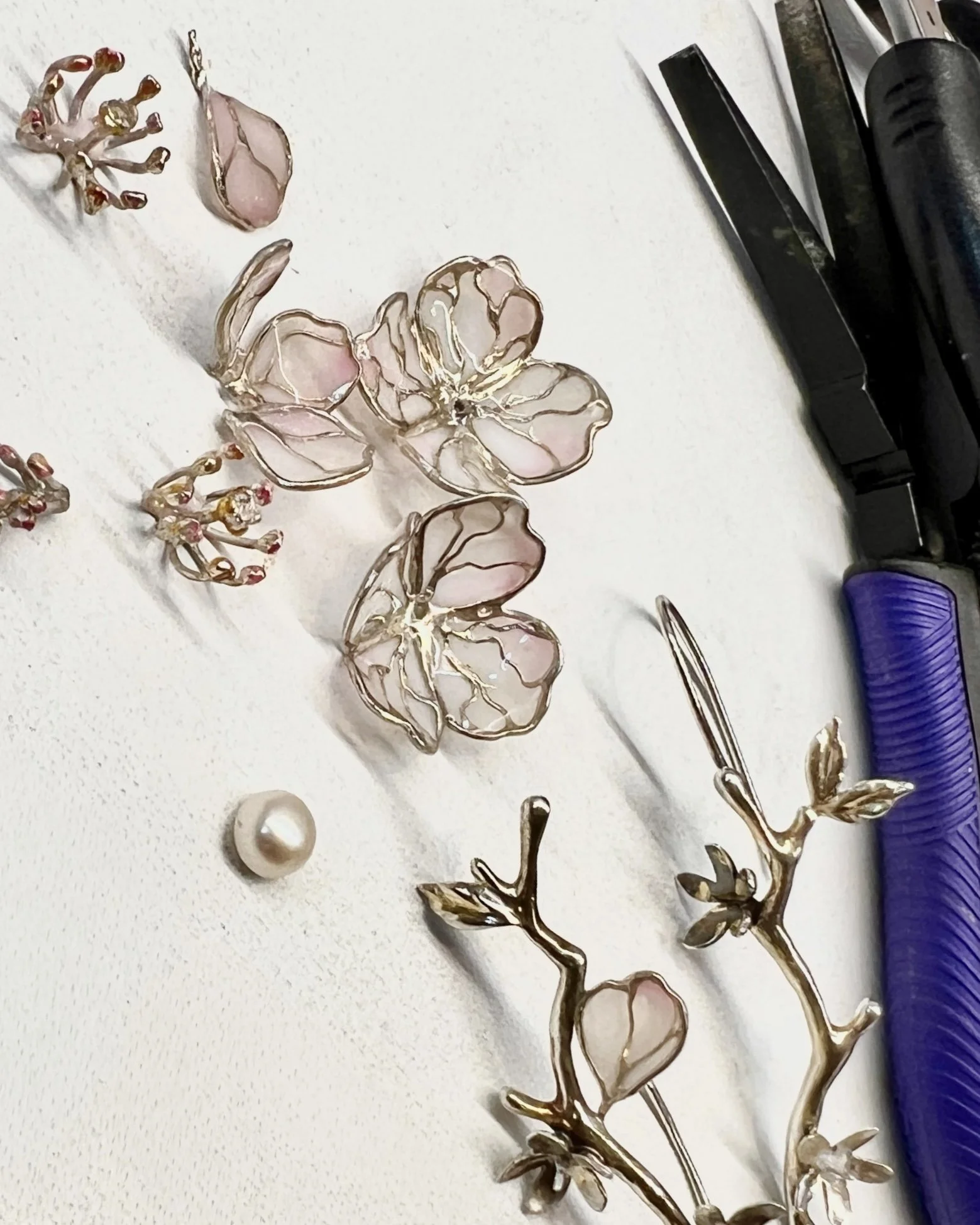 Decorative jewelry pieces, including floral and branch designs made from pink and gold-colored metal with some pearl accents, on a white surface alongside tools and a spool of purple thread.