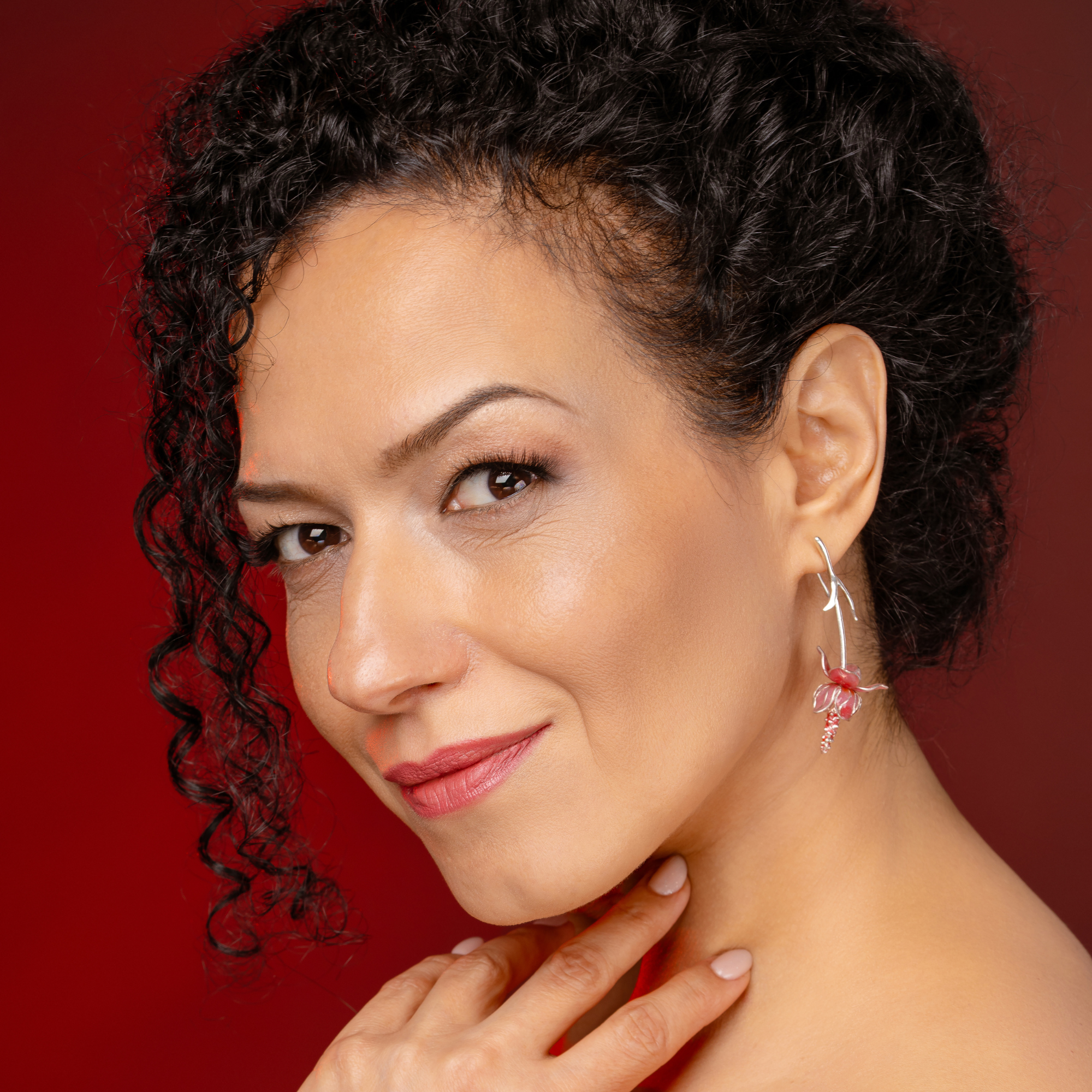 Close-up of a woman's face with curly black hair, wearing earrings, and touching her shoulder, against a red background.