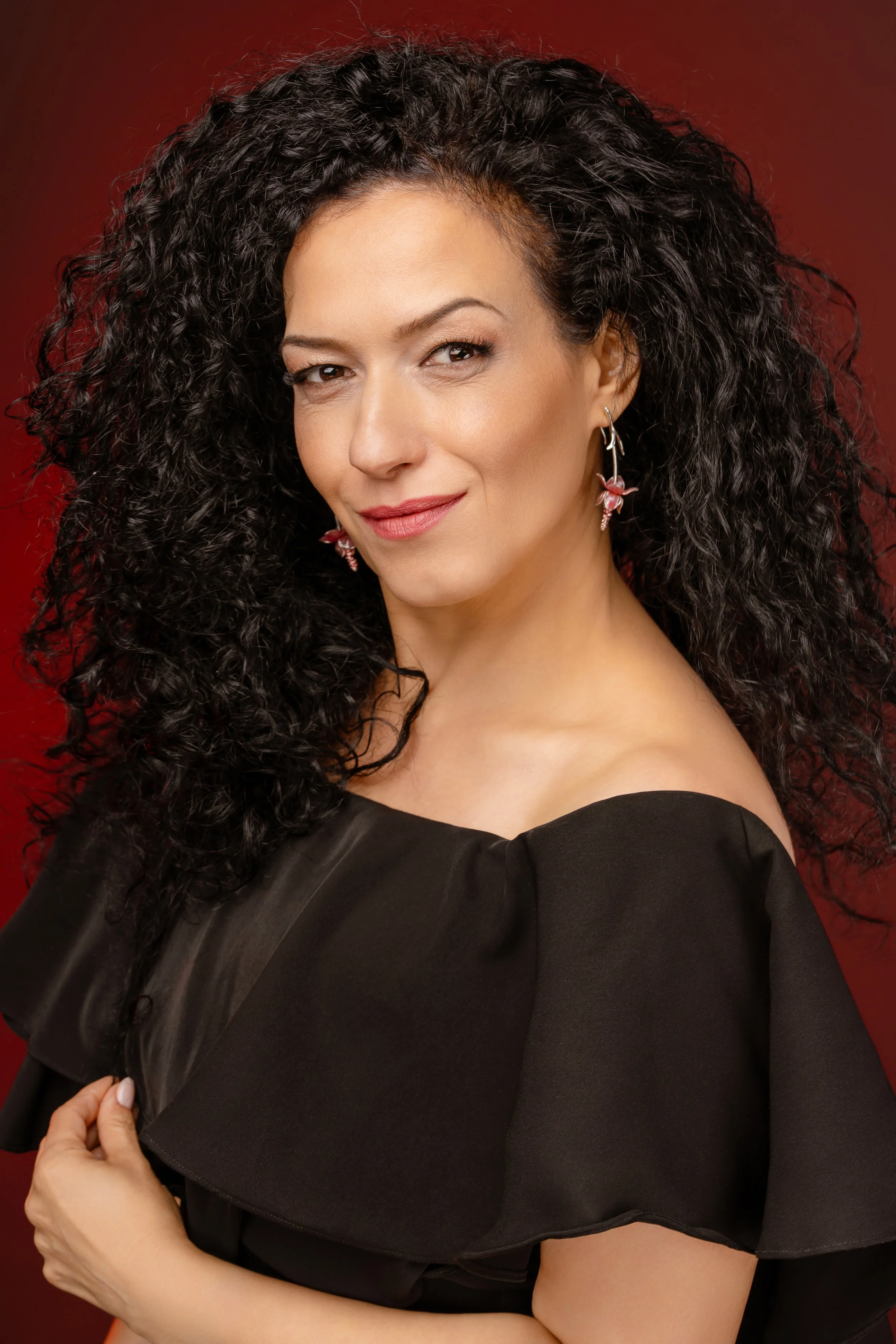 A woman with long, curly black hair winks and smiles at the camera. She wears pink flower earrings and a black dress with a ruffled neckline. The background is a deep red.