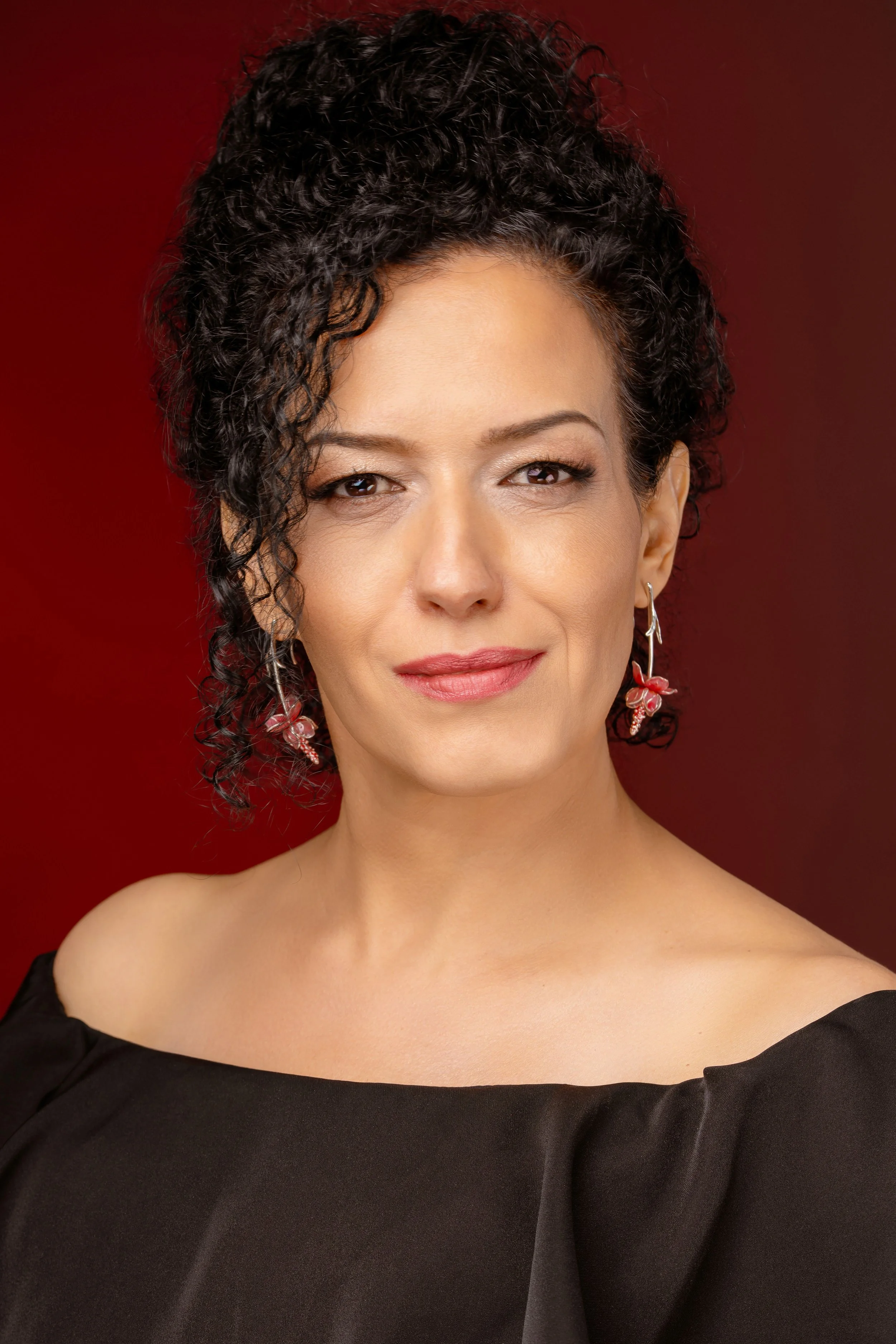 A woman with fair skin, curly black hair, and makeup, wearing earrings and a black off-the-shoulder top, poses in front of a red background.