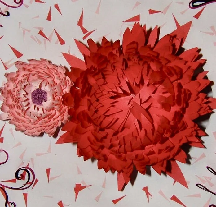 Two paper flowers, one large red and one smaller pink, on a white surface with red confetti and black and pink curls.