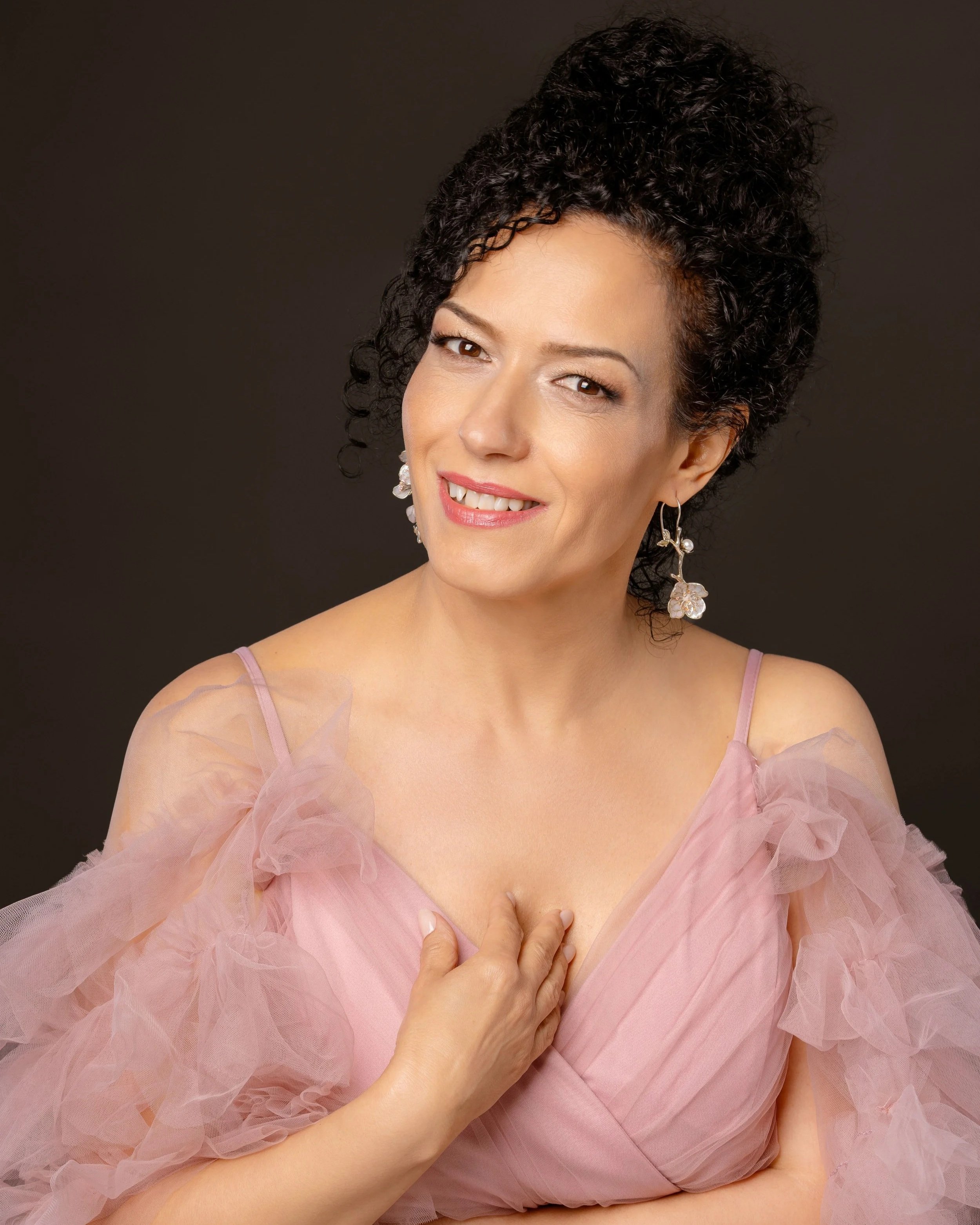 A woman with curly dark hair, wearing earrings, a pink dress with sheer ruffled fabric, and making a gesture with her hand on her chest against a dark background.