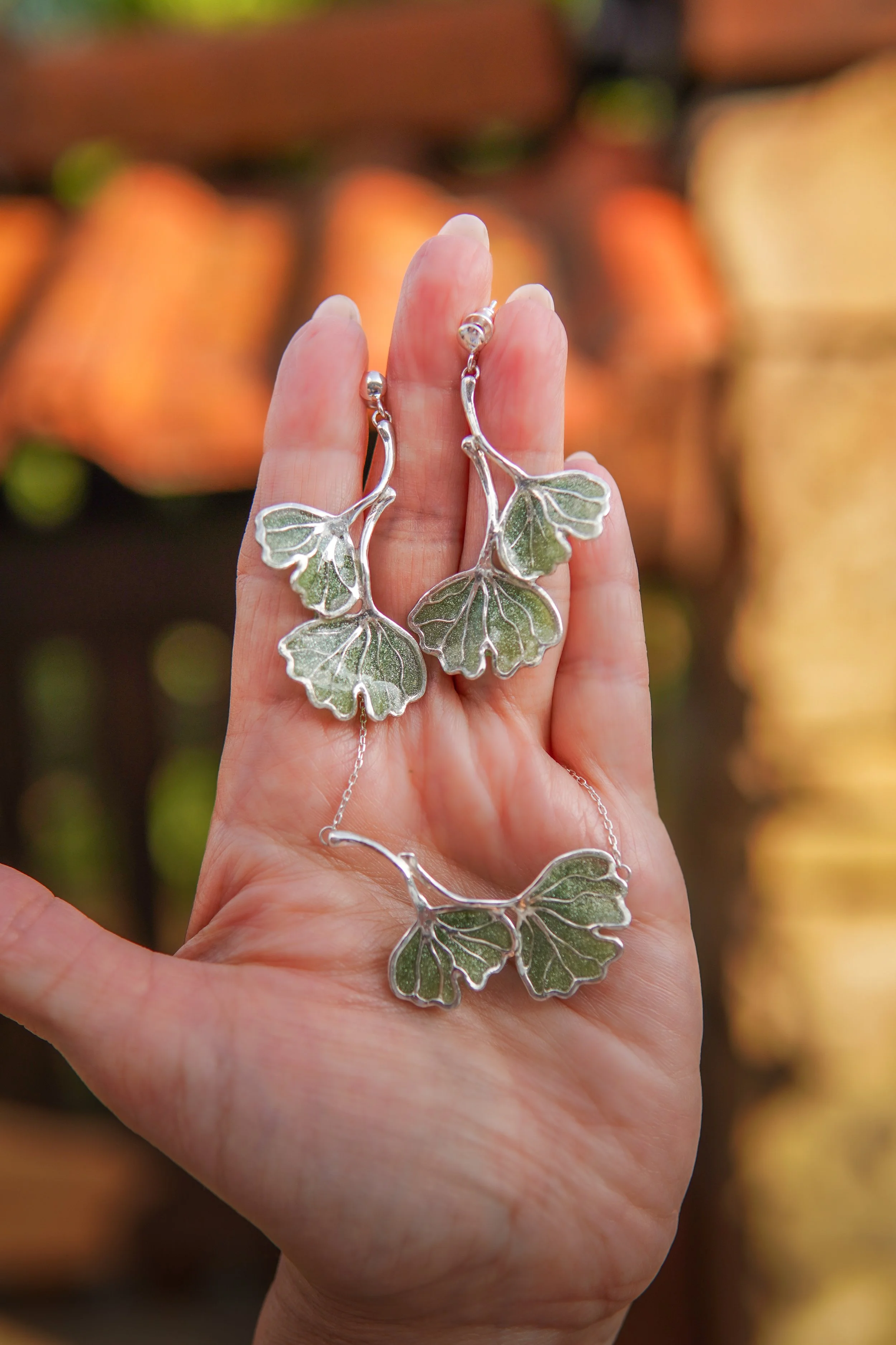 Hand holding a silver necklace with leaf-shaped pendants in front of a blurred background of orange and yellow pumpkins.