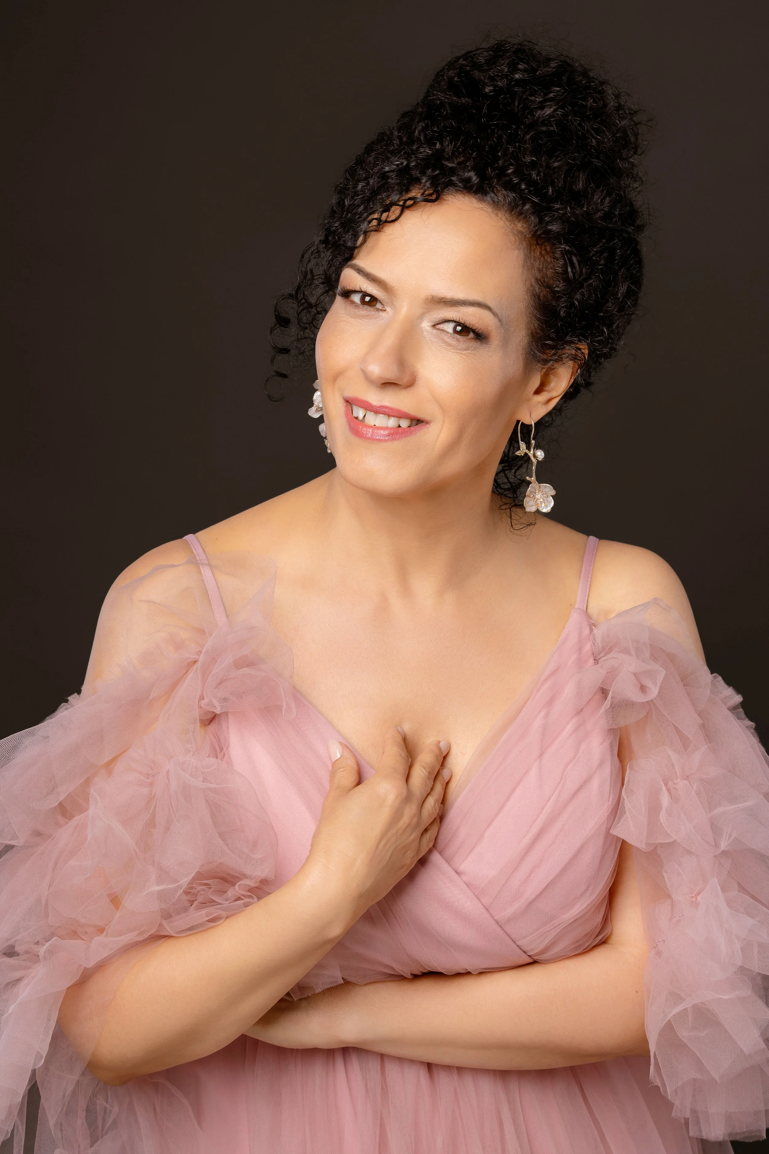 A woman wearing a pink dress with tulle ruffles, smiling and touching her chest with one hand, against a dark background.