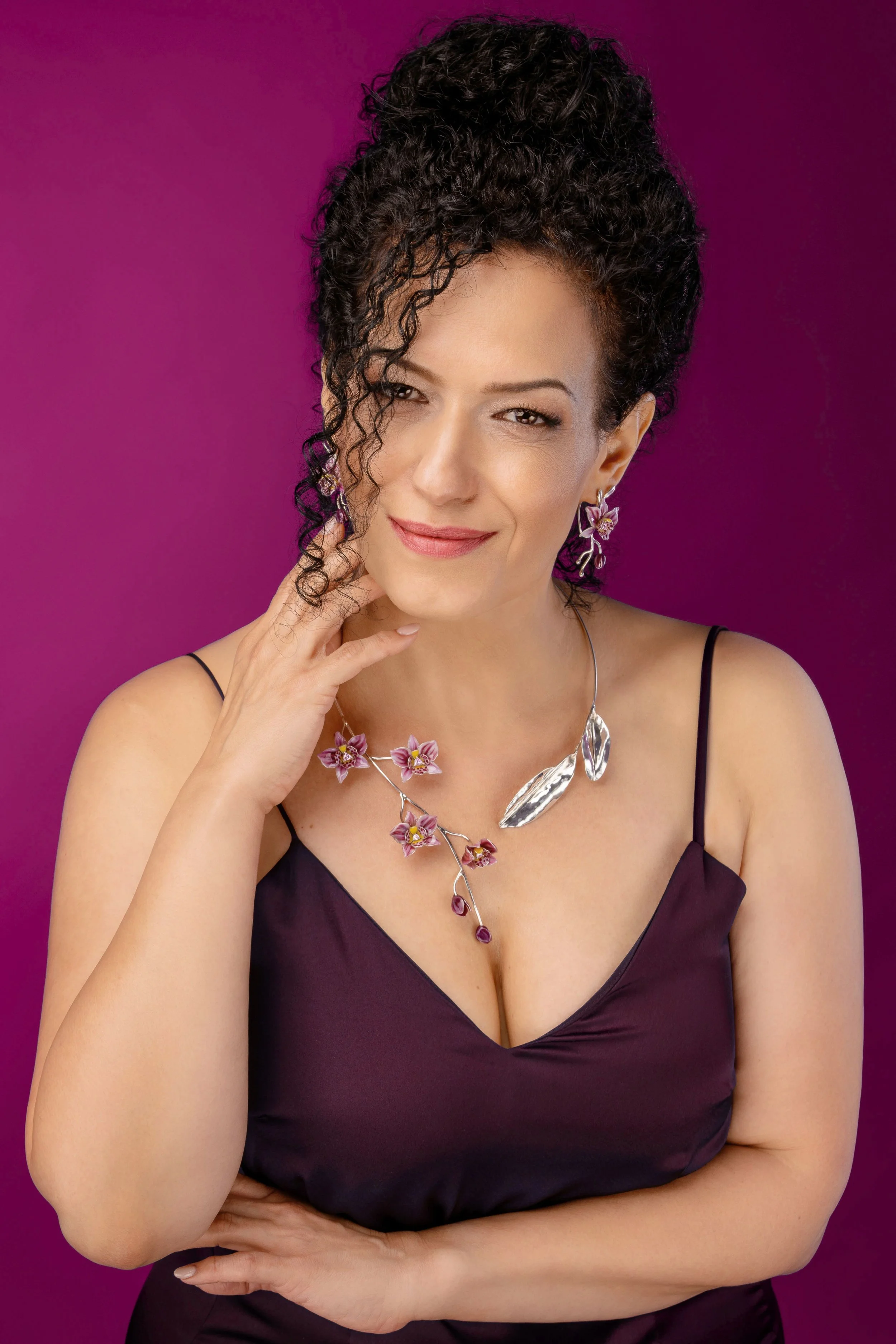 A woman in a black dress poses with a slight smile, touching her curly hair, against a purple background, wearing floral earrings and a matching necklace.
