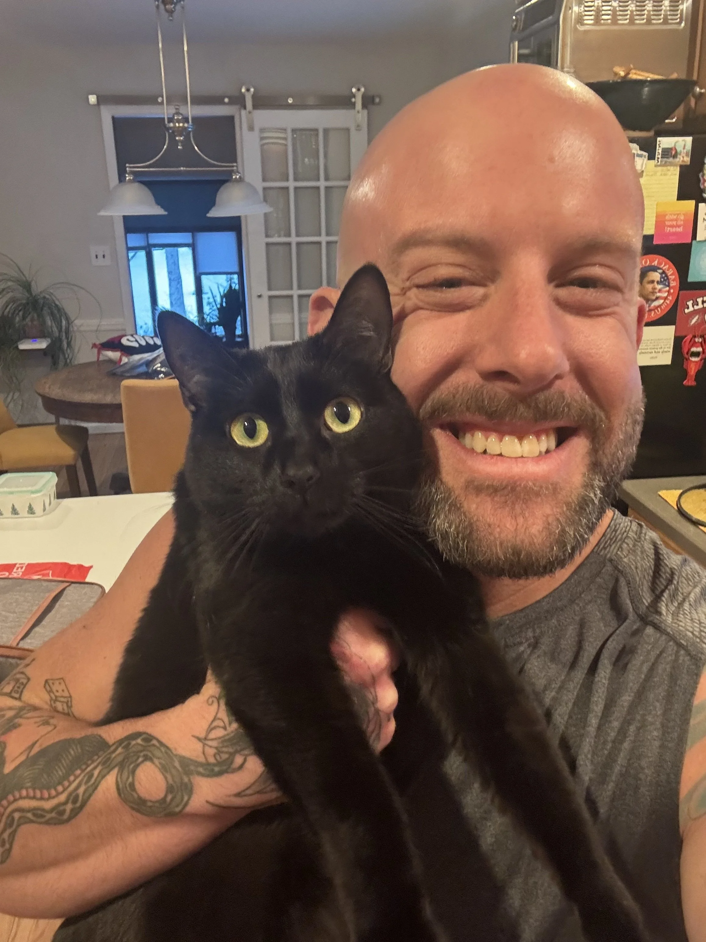A smiling man with a beard holding a black cat with yellow eyes in a kitchen.