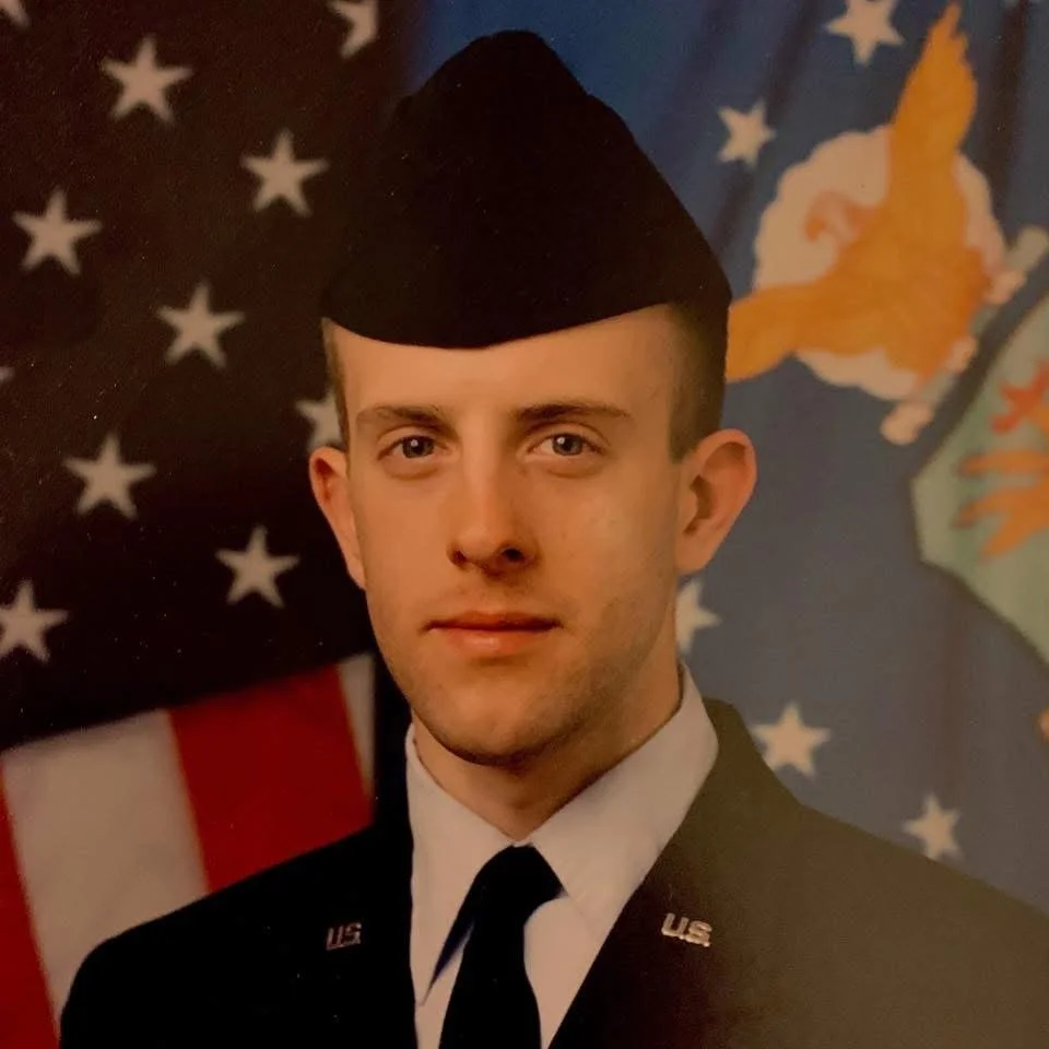 A young man in a U.S. Army dress uniform with a garrison cap, standing in front of U.S. flags.
