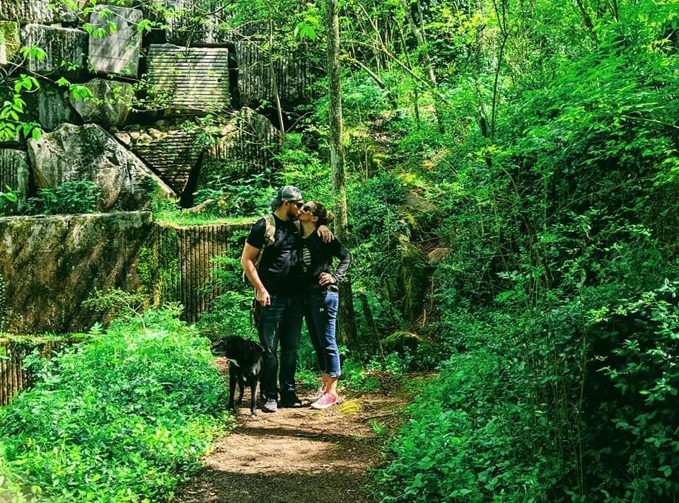 Two people, a man and a woman, hugging each other on a forest trail with a black dog nearby, surrounded by lush green trees and plants.