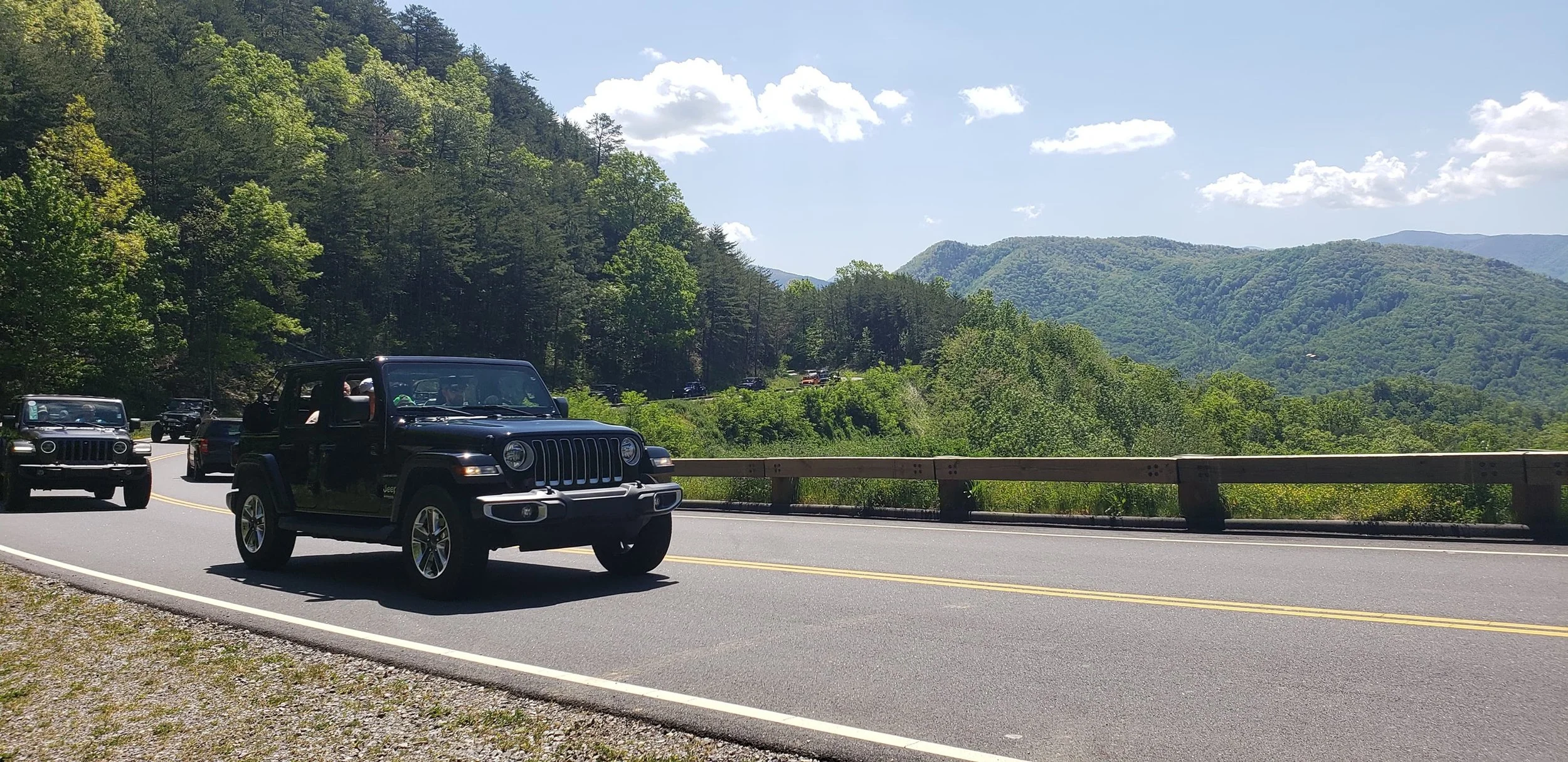 Black Jeep SUV driving on a winding mountain road with a guardrail, surrounded by lush green trees and mountains under a partly cloudy sky.