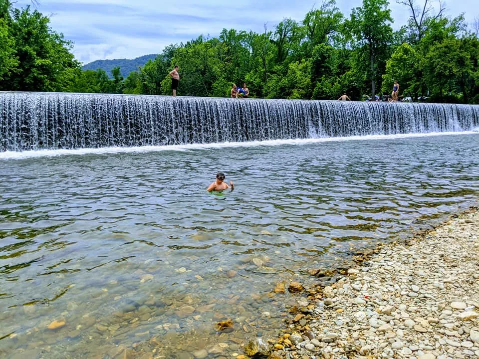 People enjoying a day outdoors at a waterfall with a rocky shore and green trees in the background. One person is swimming in the water, while others are standing or sitting on the water's edge or a small ledge.