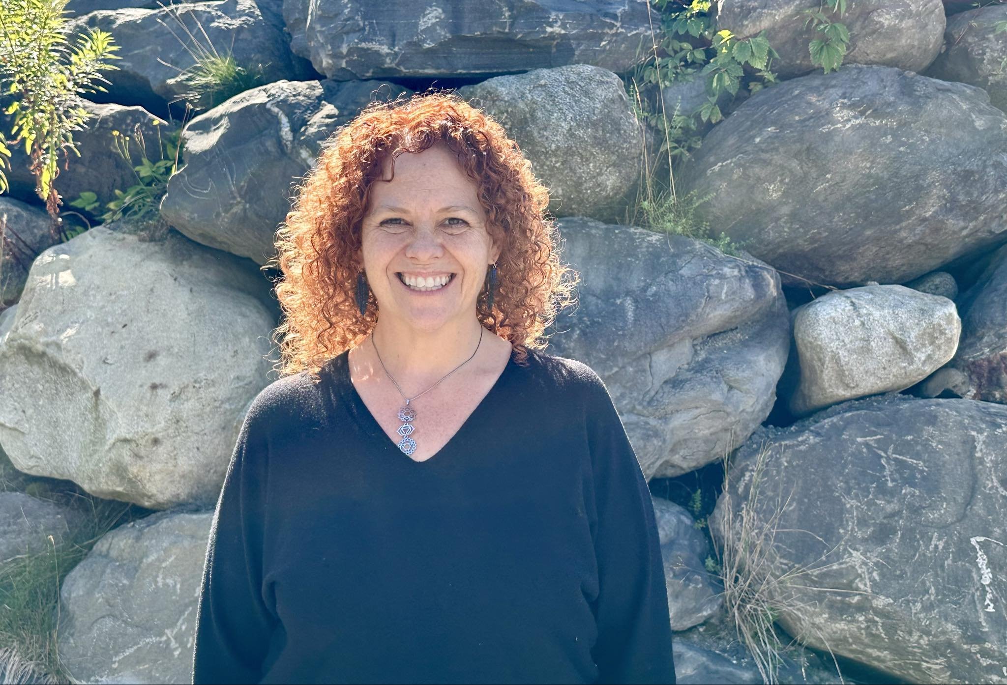 A woman with curly red hair smiling, standing outdoors in front of a large rock formation with green plants.