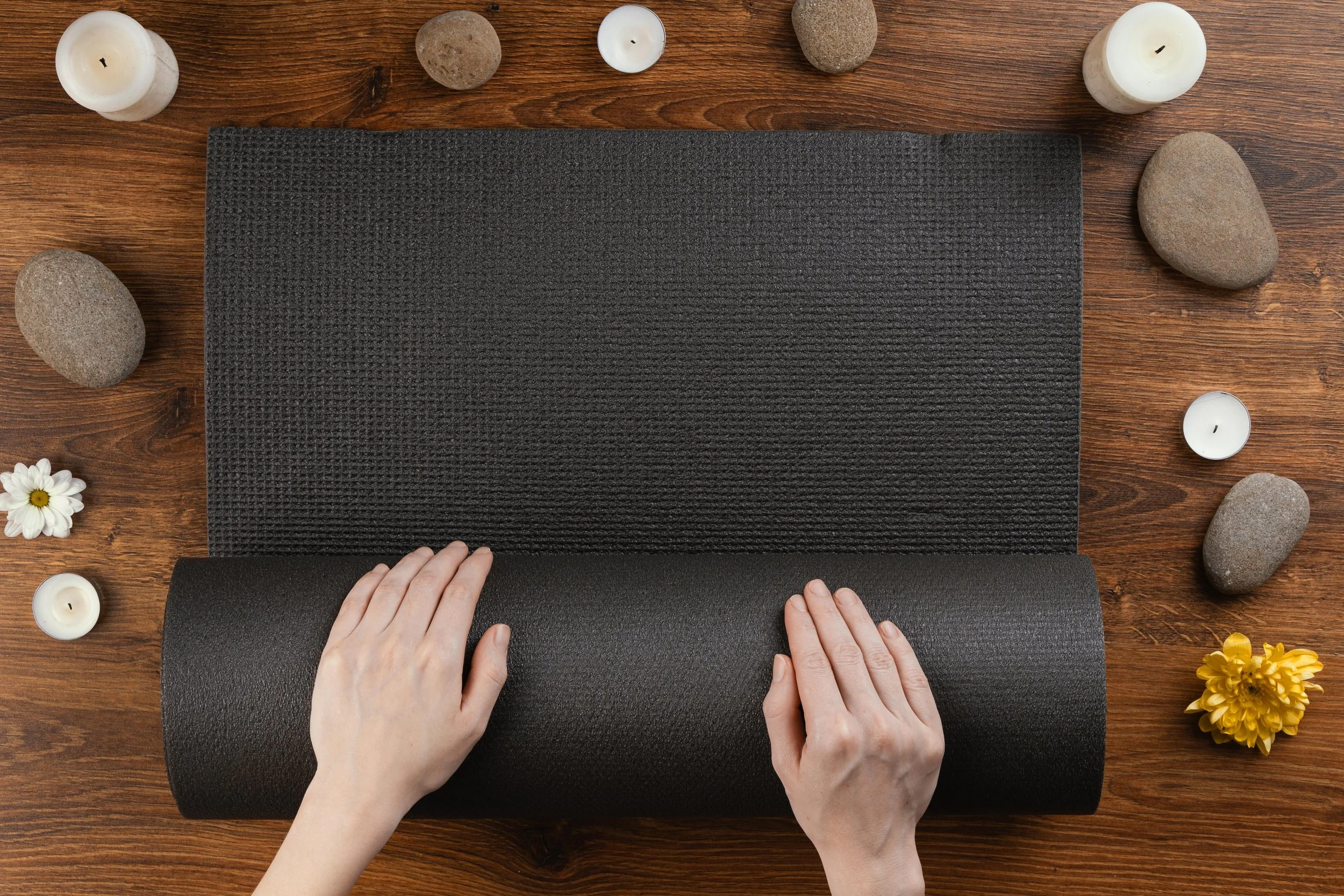 Person handling black yoga mats on a wooden surface surrounded by candles, stones, flowers, and a yellow flower arrangement.