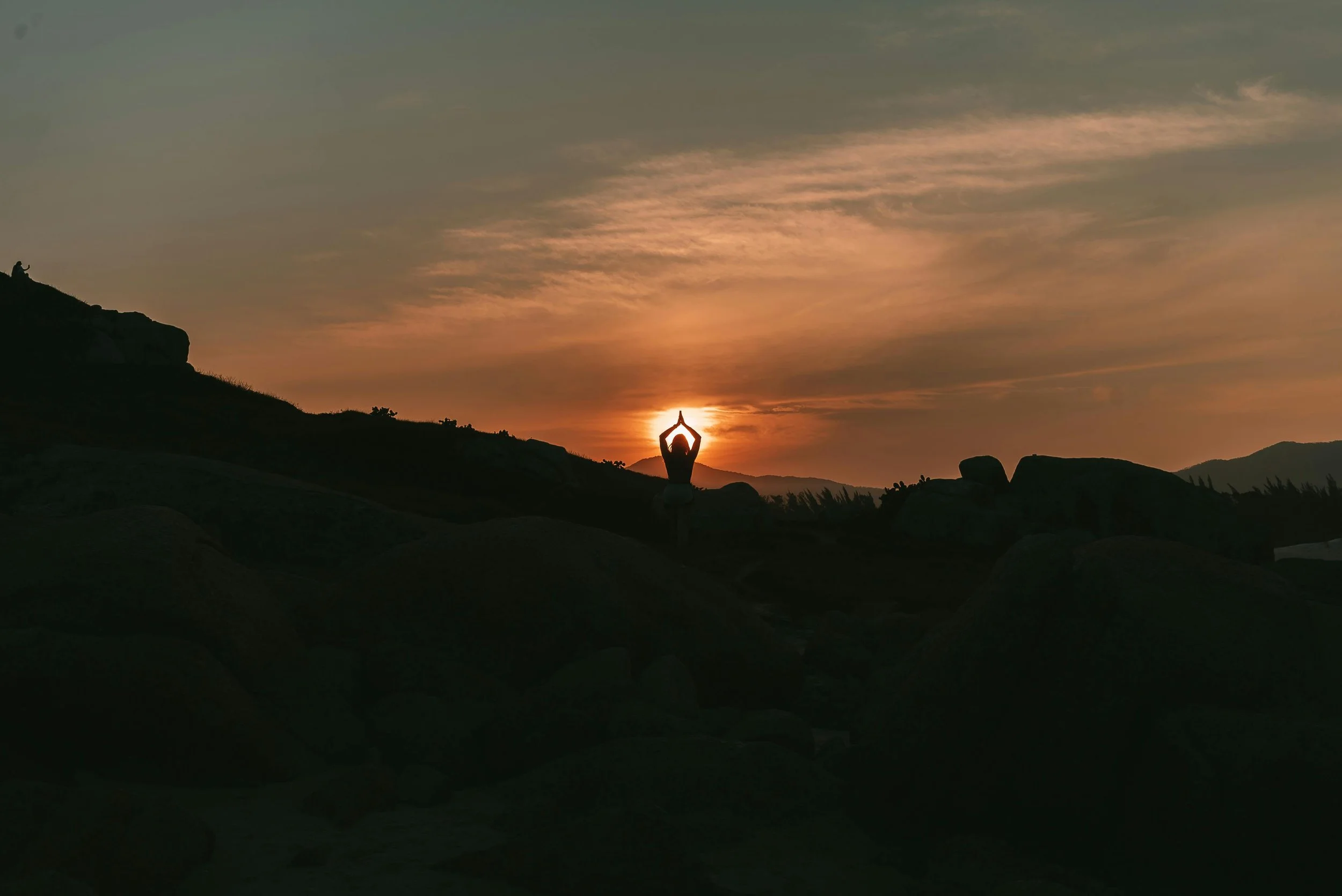 Person practicing yoga in a seated meditation pose at sunset in a natural landscape with rocks and hills.