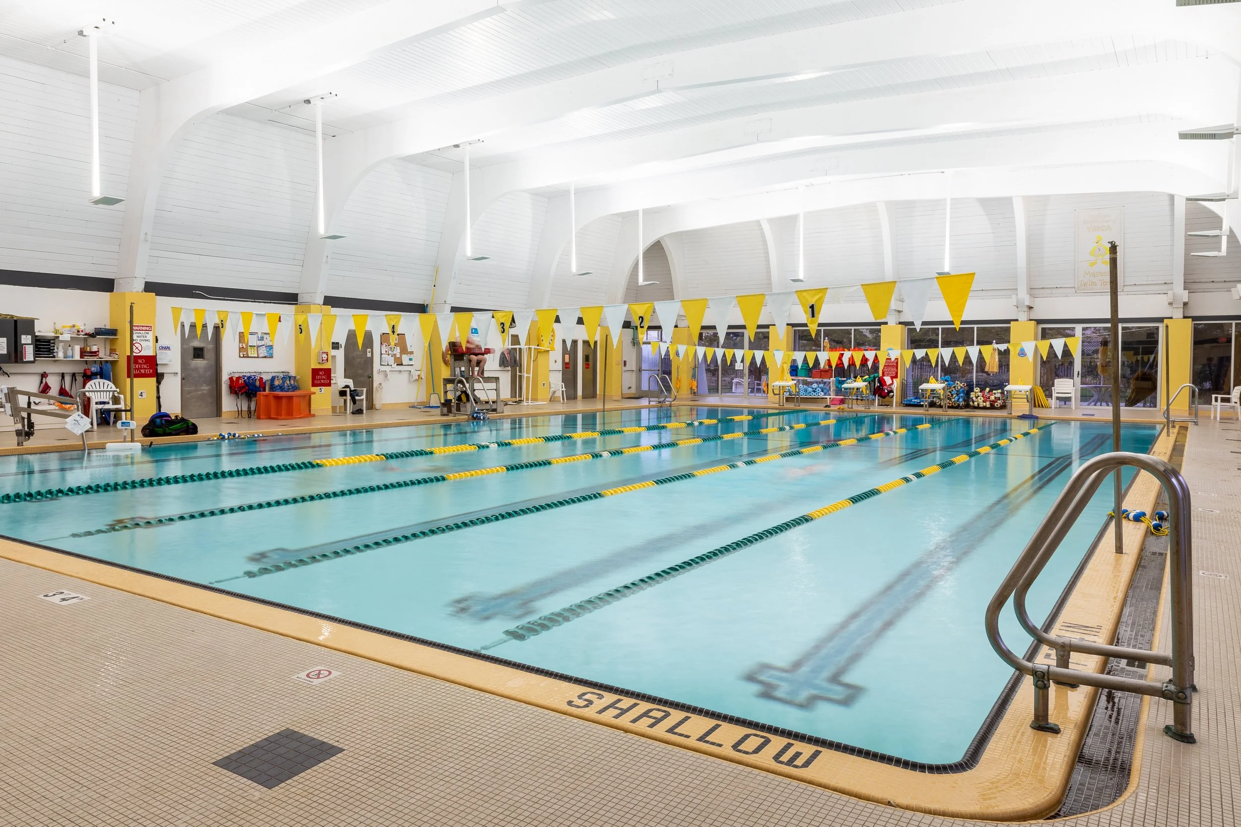 Indoor swim pool area with yellow and white lane dividers, starting blocks, and various equipment along the walls, under a white arched ceiling.