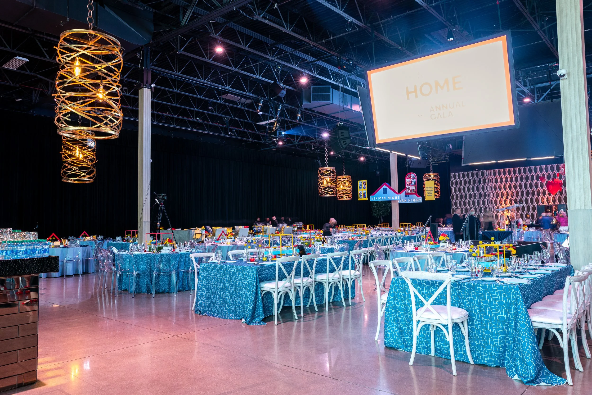 Event hall set up for a dinner or gala with tables decorated with blue patterned tablecloths and centerpieces, hanging modern light fixtures, a large screen displaying "HOME ANNUAL GALA," and a decorative partition with red heart accents.