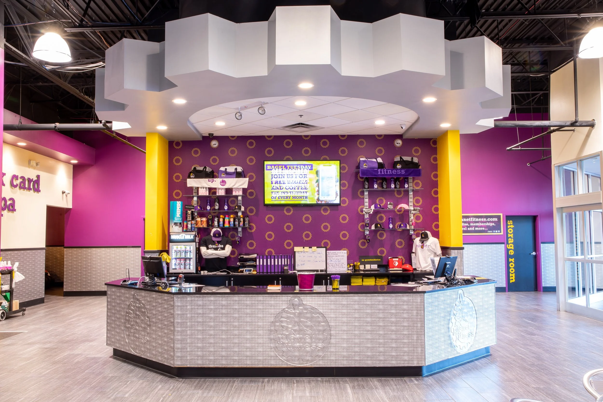 Inside a fitness store with purple and yellow walls, a counter with purple items, and a display shelf with fitness gear. A digital screen is mounted on the wall, and the ceiling has white panels with embedded lights.