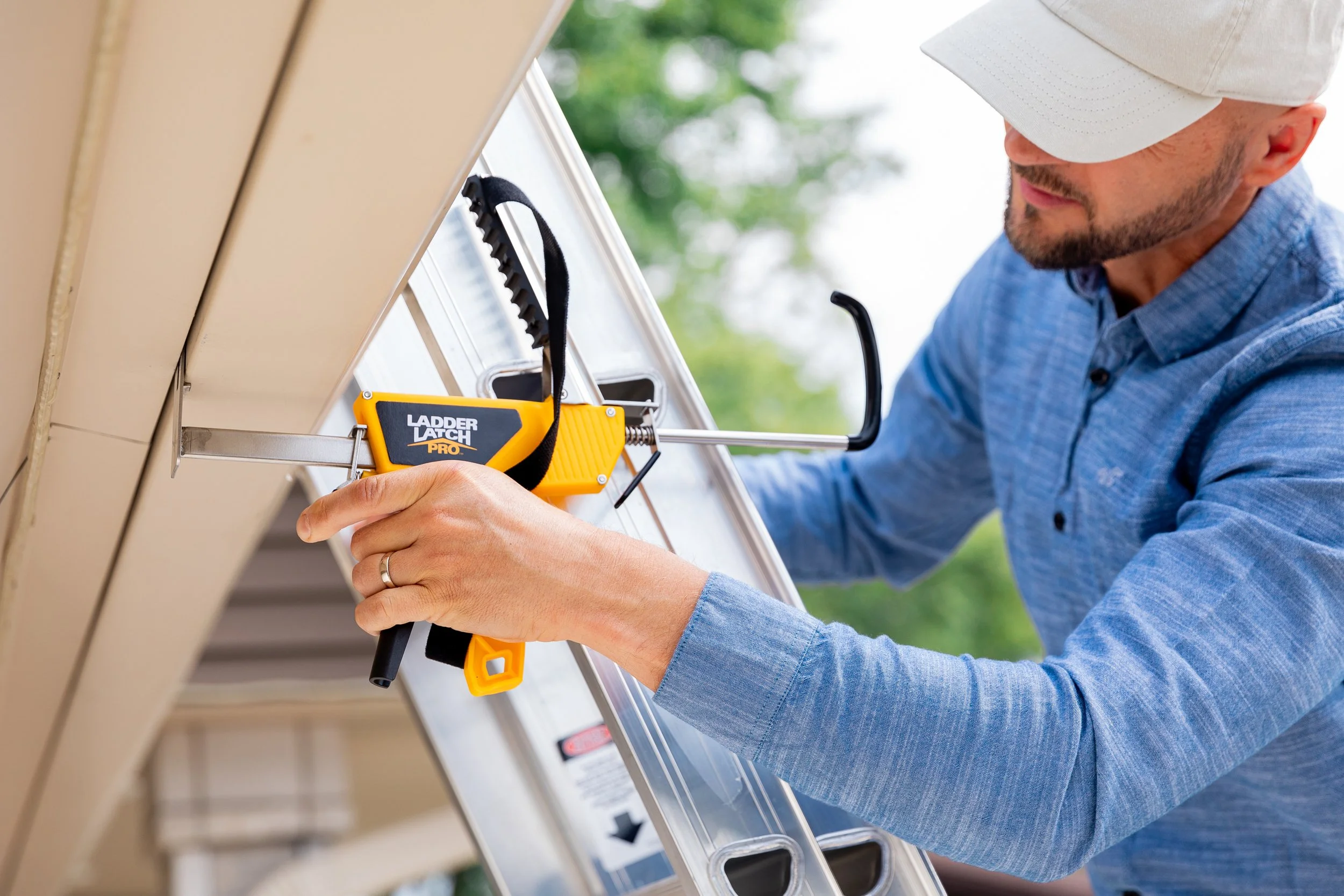 A man wearing a light-colored cap and a blue shirt using a yellow and black ladder latch pro to secure a ladder to a house exterior.