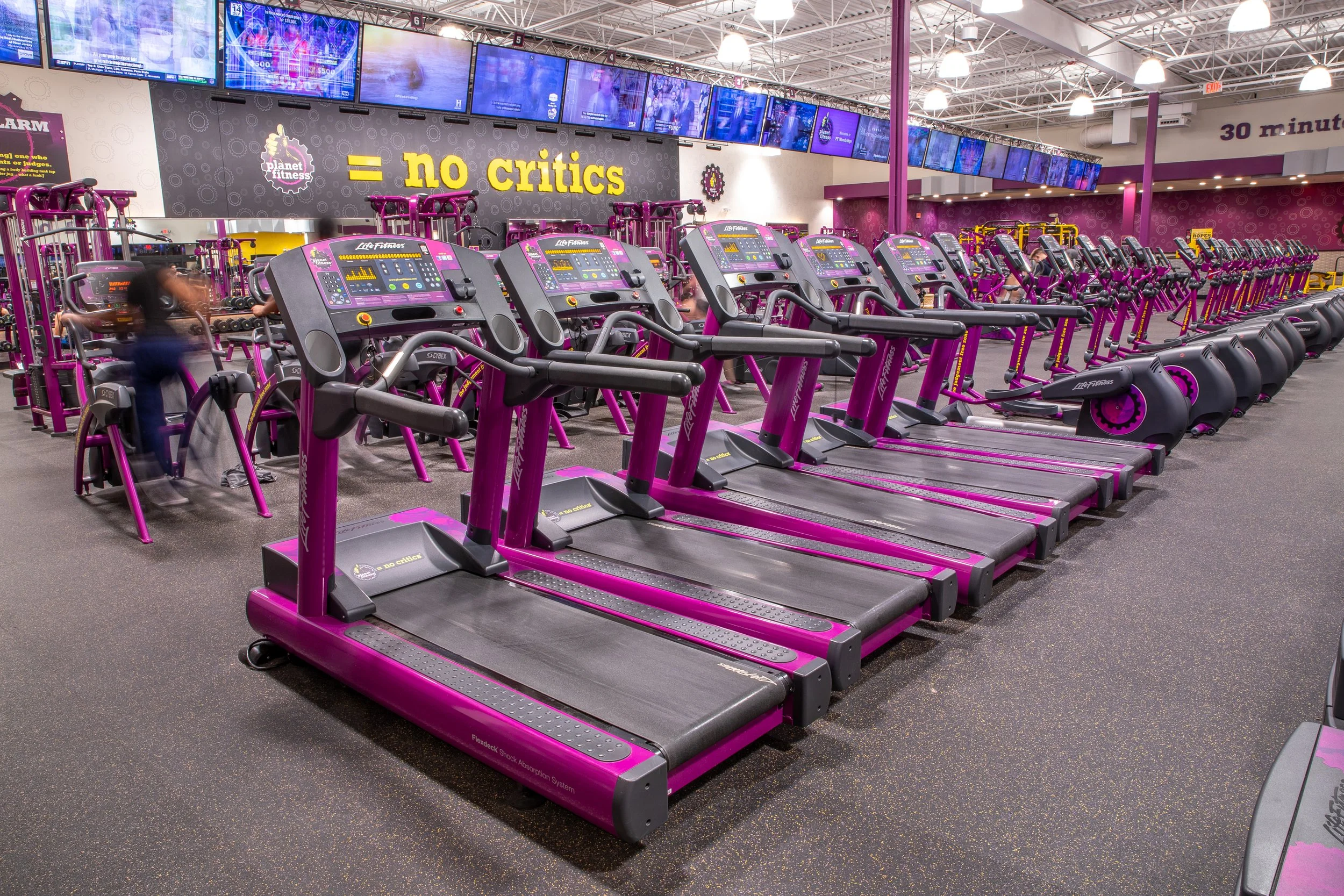 Row of purple treadmill exercise machines in a gym with multiple large screens displaying various videos and information in the background. The gym has a high ceiling with white lights and purple-colored support beams.