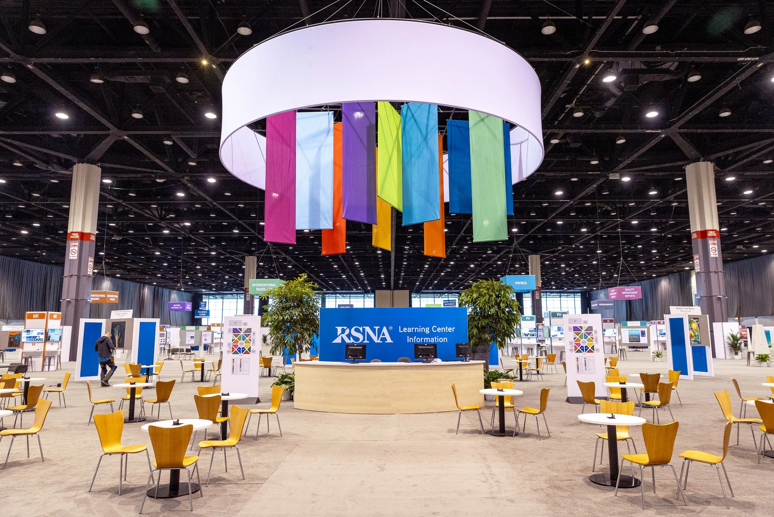An indoor convention center with a large hanging circular display featuring colorful vertical banners. The center has a reception desk labeled RSNA Learning Center with several informational booths and numerous yellow chairs and tables scattered arou