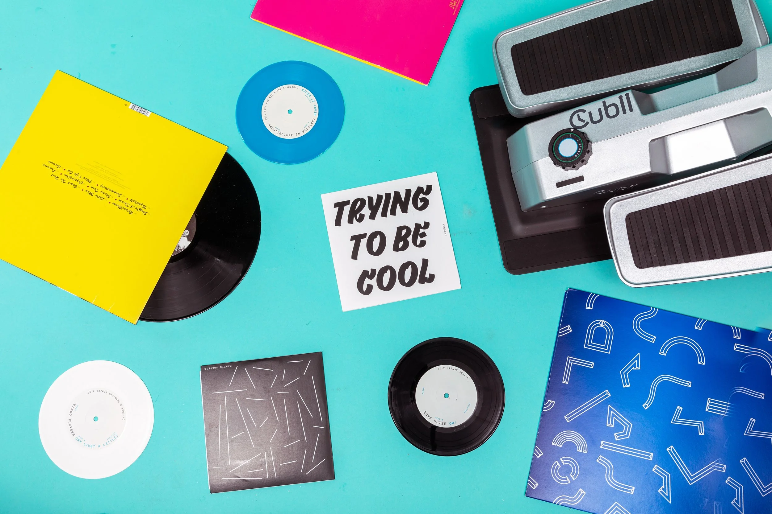 Flat lay of vinyl records, colorful books, and a small machine on a turquoise surface with a sticker that says 'TRYING TO BE COOL'.