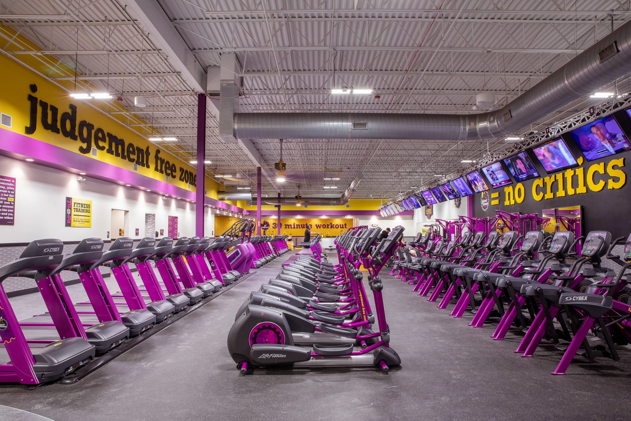 Inside a gym with rows of purple and black exercise treadmills and elliptical machines, with yellow and purple walls and multiple TV screens hanging from the ceiling.