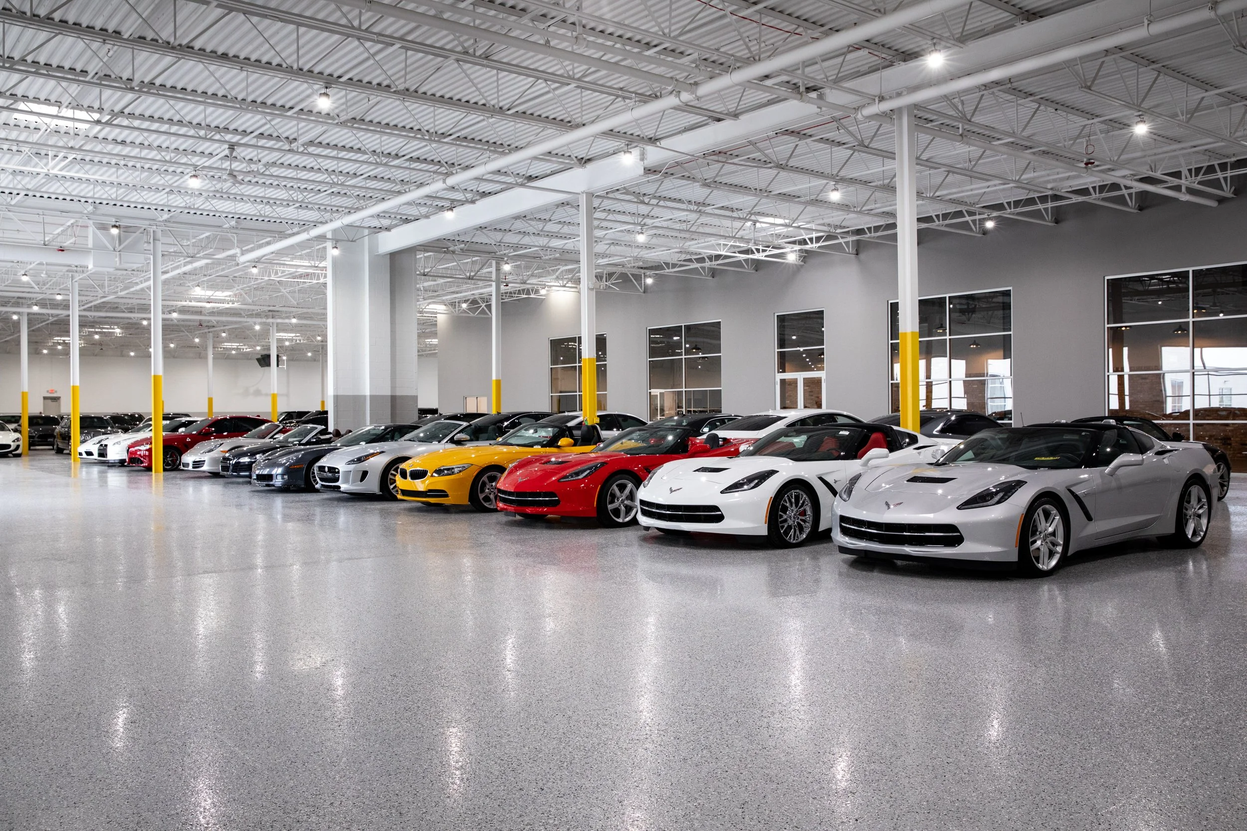 Indoor car dealership displaying various sports cars in different colors, arranged in a row in a well-lit showroom with polished concrete floors and large windows.