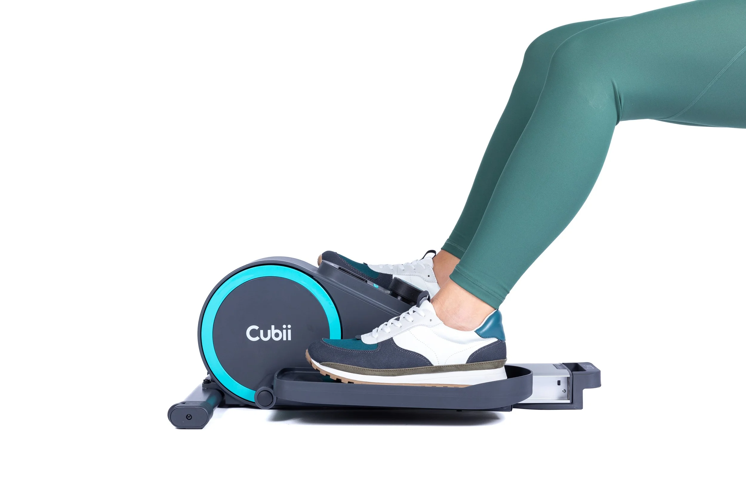 Person in teal workout leggings and white sneakers using a Cubii under-desk elliptical trainer against a white background.