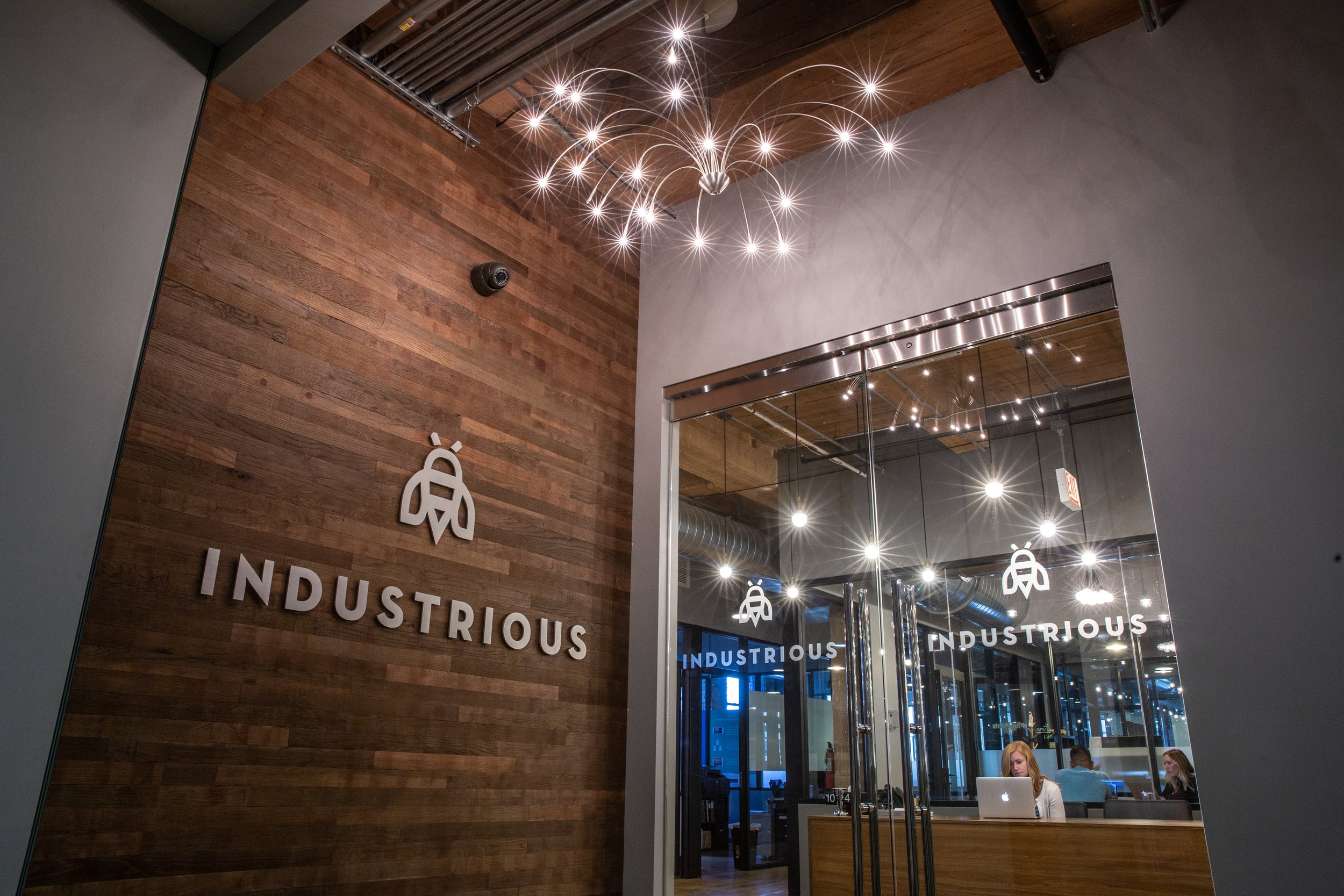 Interior view of a modern office or coworking space with a wooden wall featuring a logo and the word 'INDUSTRIOUS'. The space has a glass wall with metal framing, multiple ceiling lights creating starburst effects, and people working at desks visible