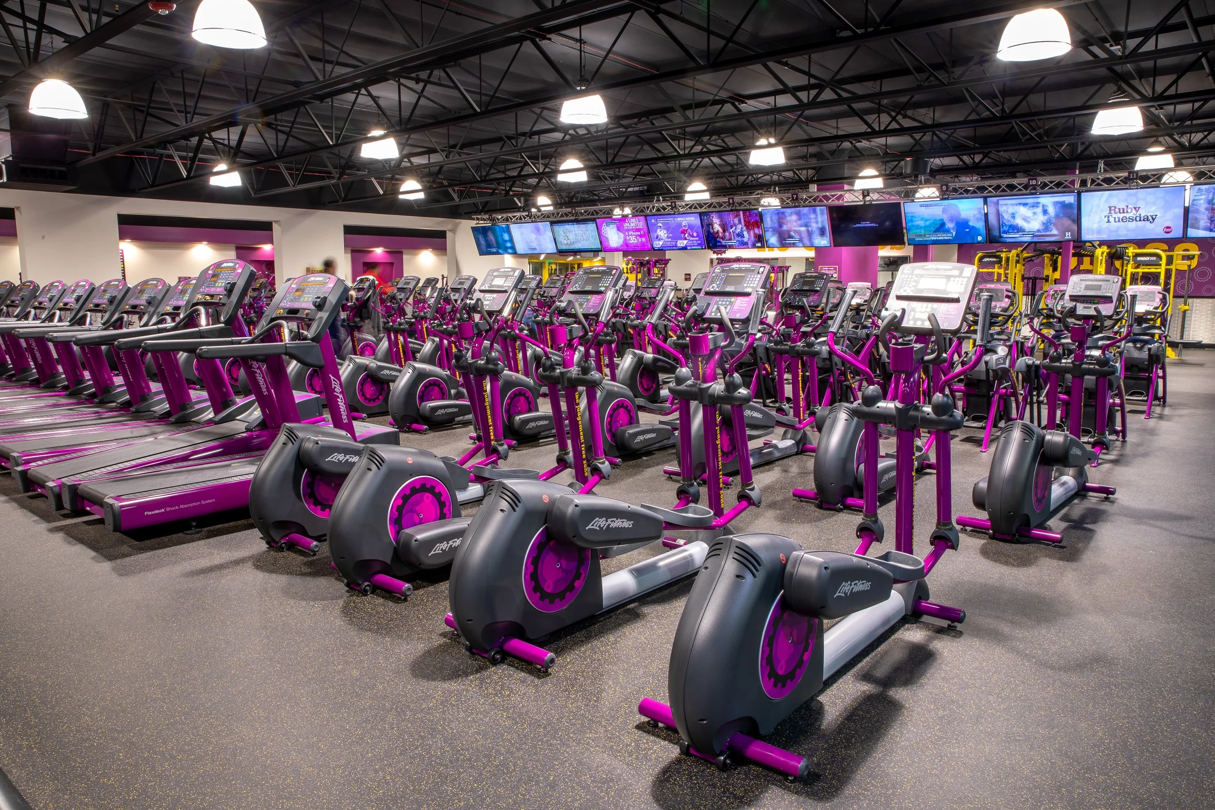 Indoor gym with rows of purple elliptical machines and treadmills, multiple digital screens on the wall, ceiling lights, and exercise equipment in the background.