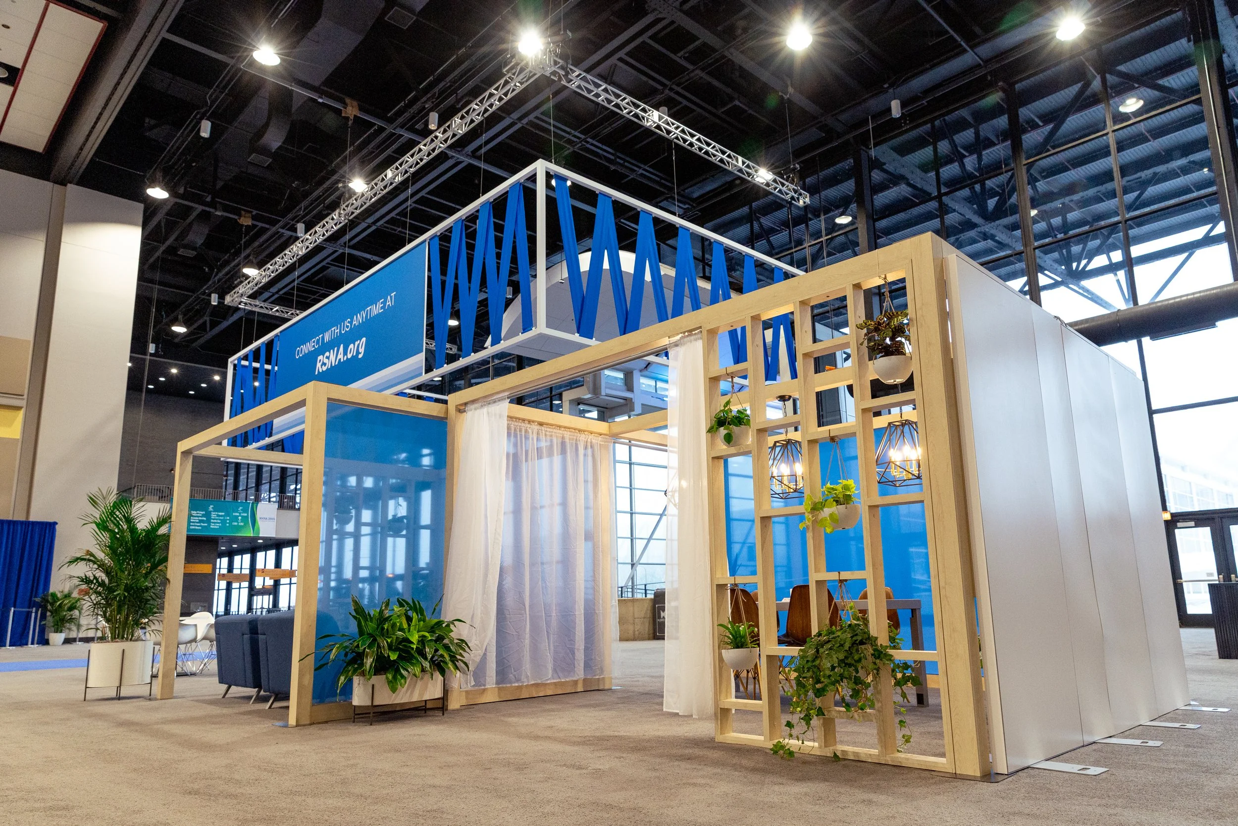 Trade show booth with wooden framework, plants, and blue accents, inside a large convention center with high ceilings, metal trusses, and large windows.