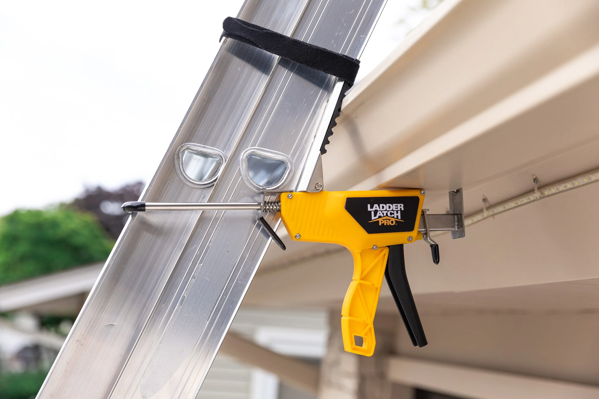 A close-up of a silver folding ladder with a black protective strap, attached to a yellow tool called Ladder Latch Pro, used for securing the ladder when extended.