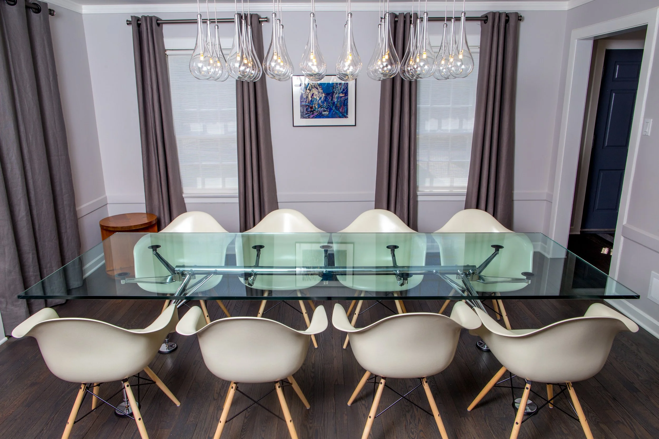 Dining room with modern glass table, cream chairs, dark hardwood floors, gray curtains, framed cityscape artwork, and a cluster of glass light fixtures hanging from the ceiling.