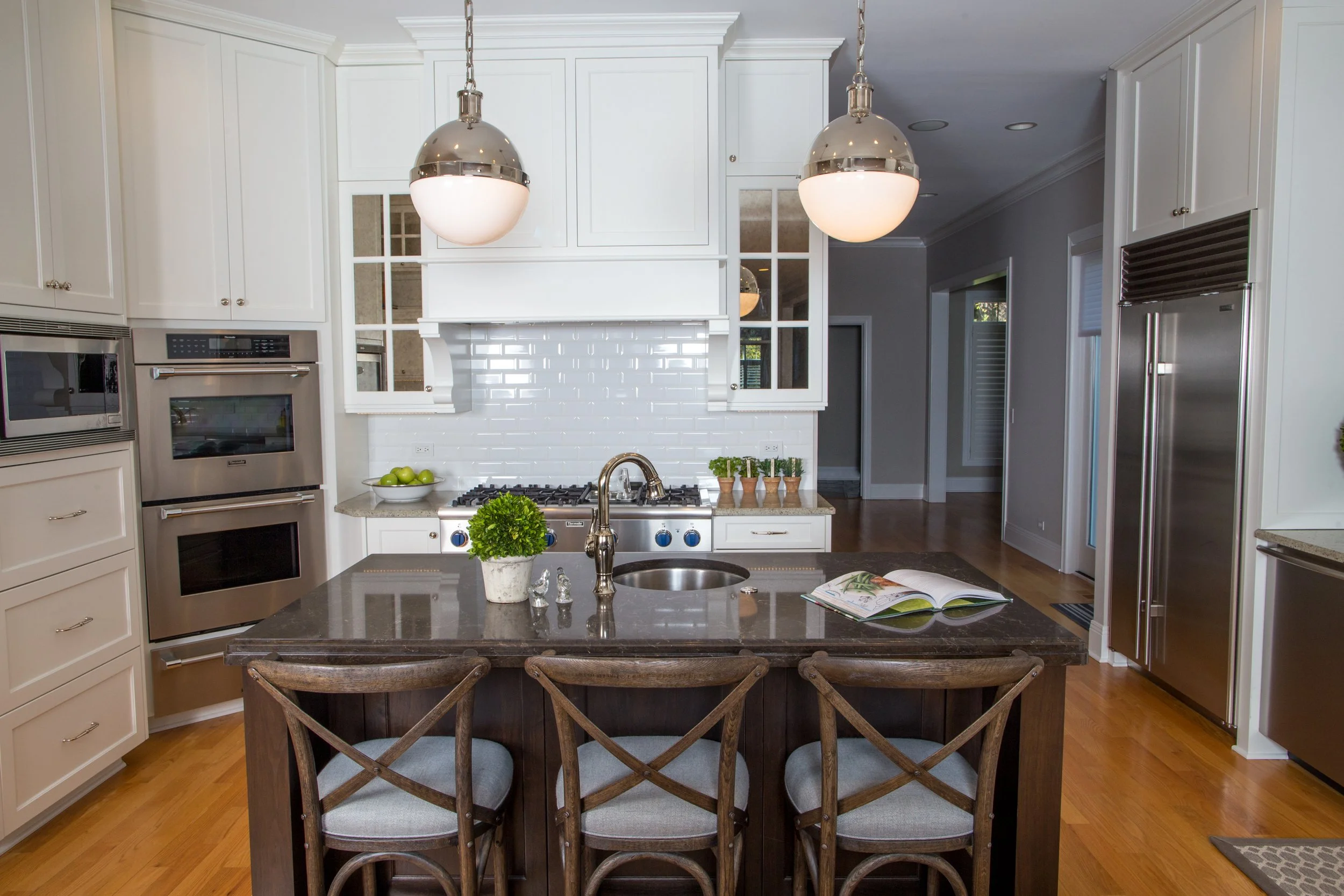 Kitchen with white cabinets, stainless steel appliances, a kitchen island with a dark wood base, a gray countertop, a small potted plant, and an open book, two large pendant lights hanging over the island, hardwood floors, and a white subway tile bac