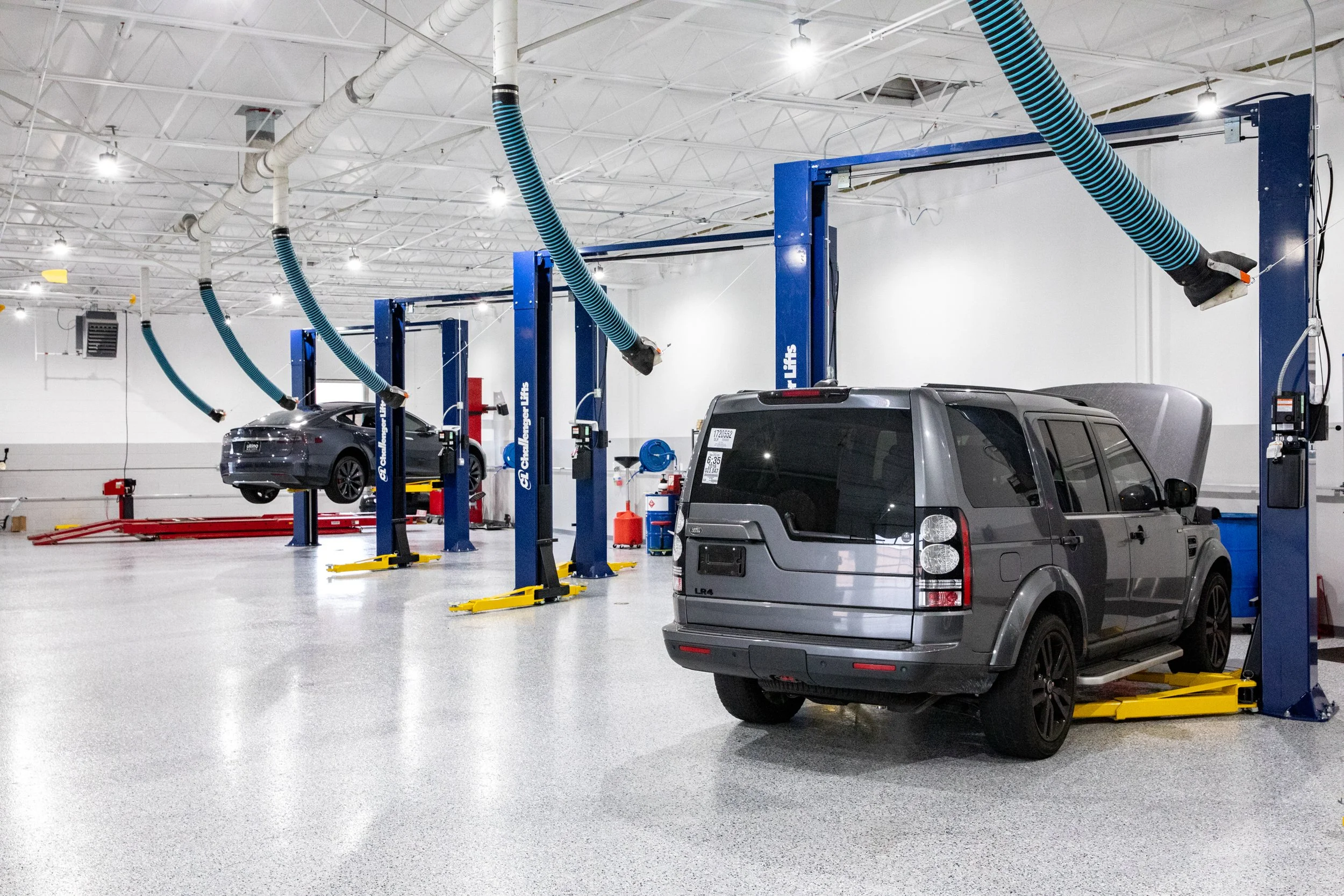 An indoor car service or repair shop with multiple vehicle lifts, a gray SUV on the floor, and a gray sedan lifted off the ground. The shop has a high ceiling with white exposed beams and industrial lighting.