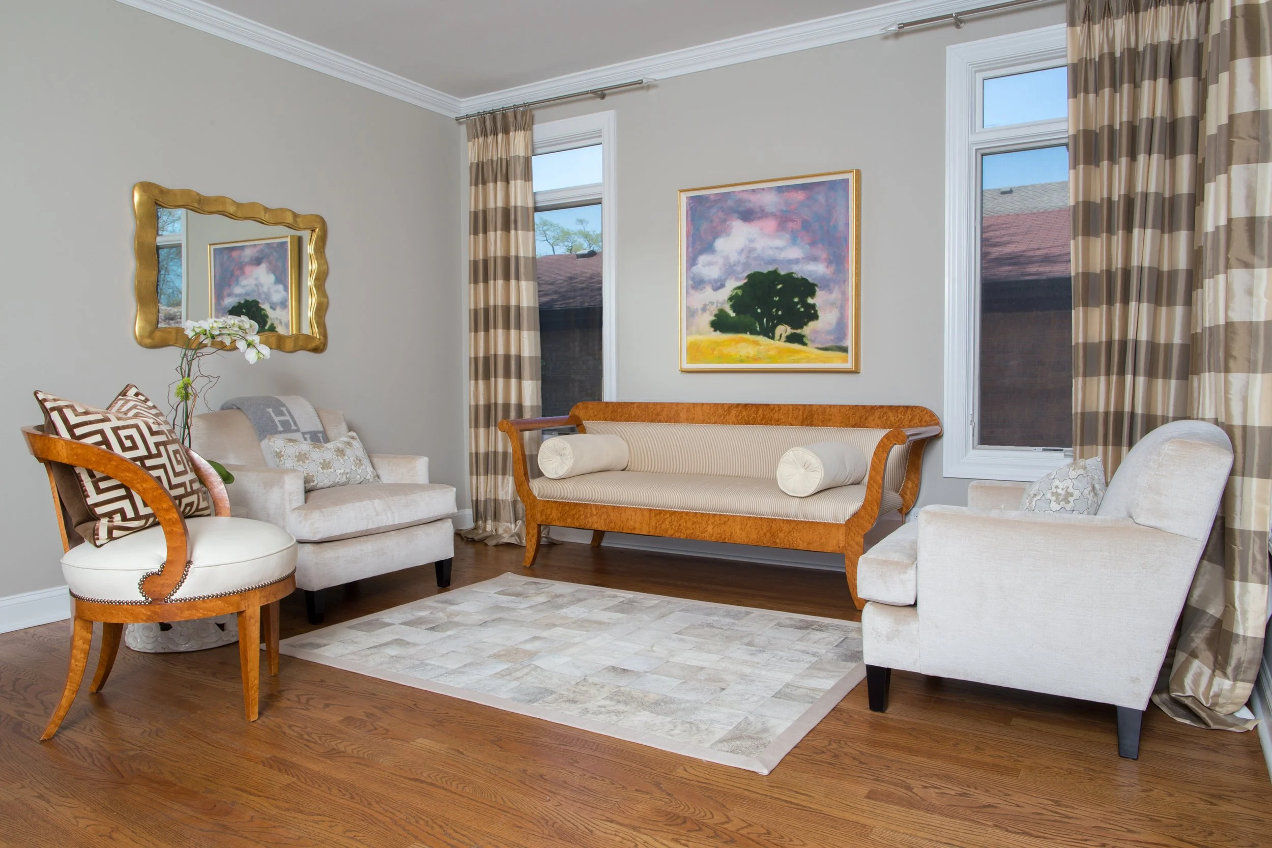 Living room with cream-colored couch and armchair, wooden bench, patterned curtains, framed painting of a tree and sky, mirror, area rug, hardwood floor, open window with blue sky.