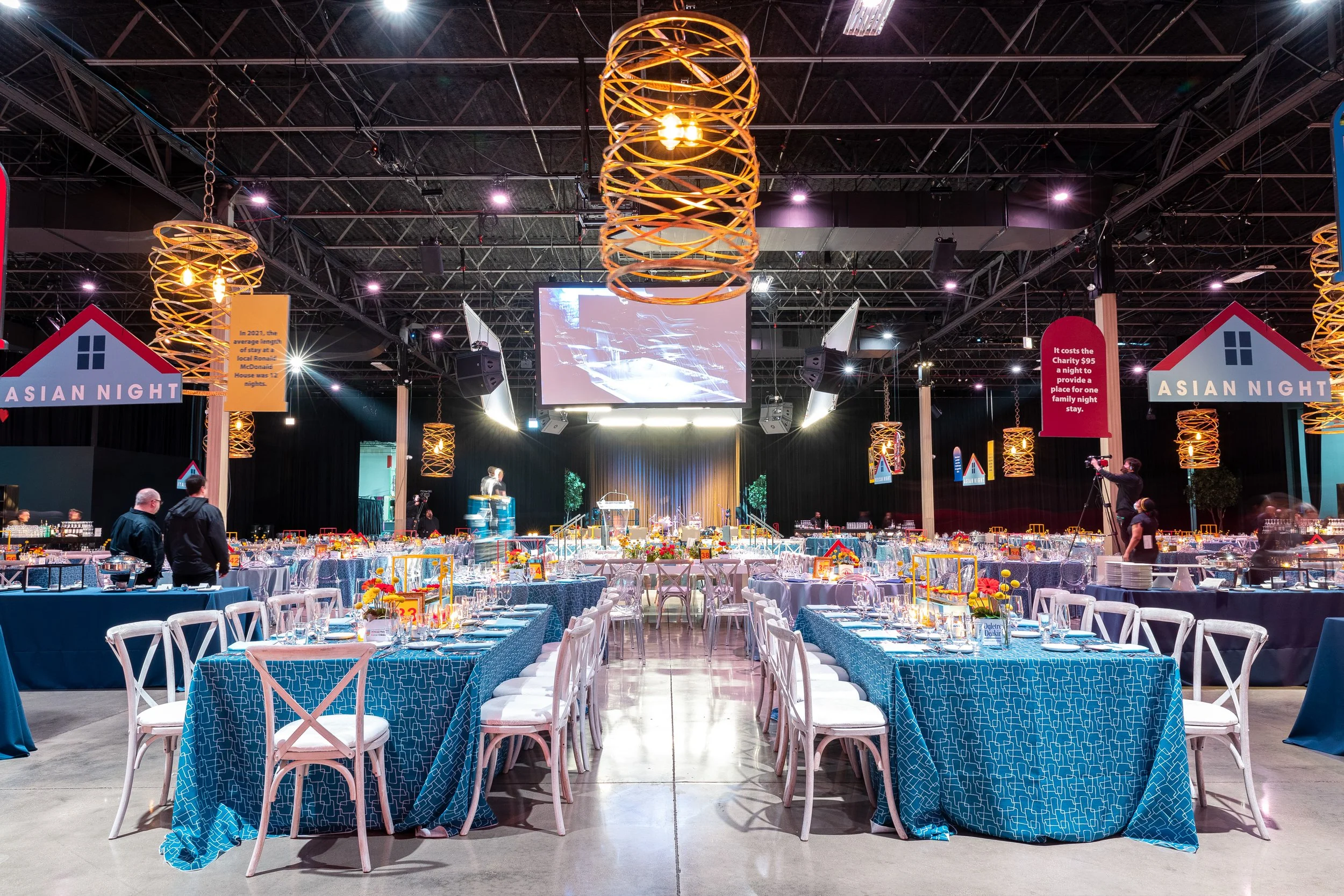 Indoor event space set for a banquet with round tables covered in blue tablecloths and white chairs, decorated with yellow flowers. Large round hanging light fixtures are suspended from the ceiling, and a stage with a large screen is visible at the f
