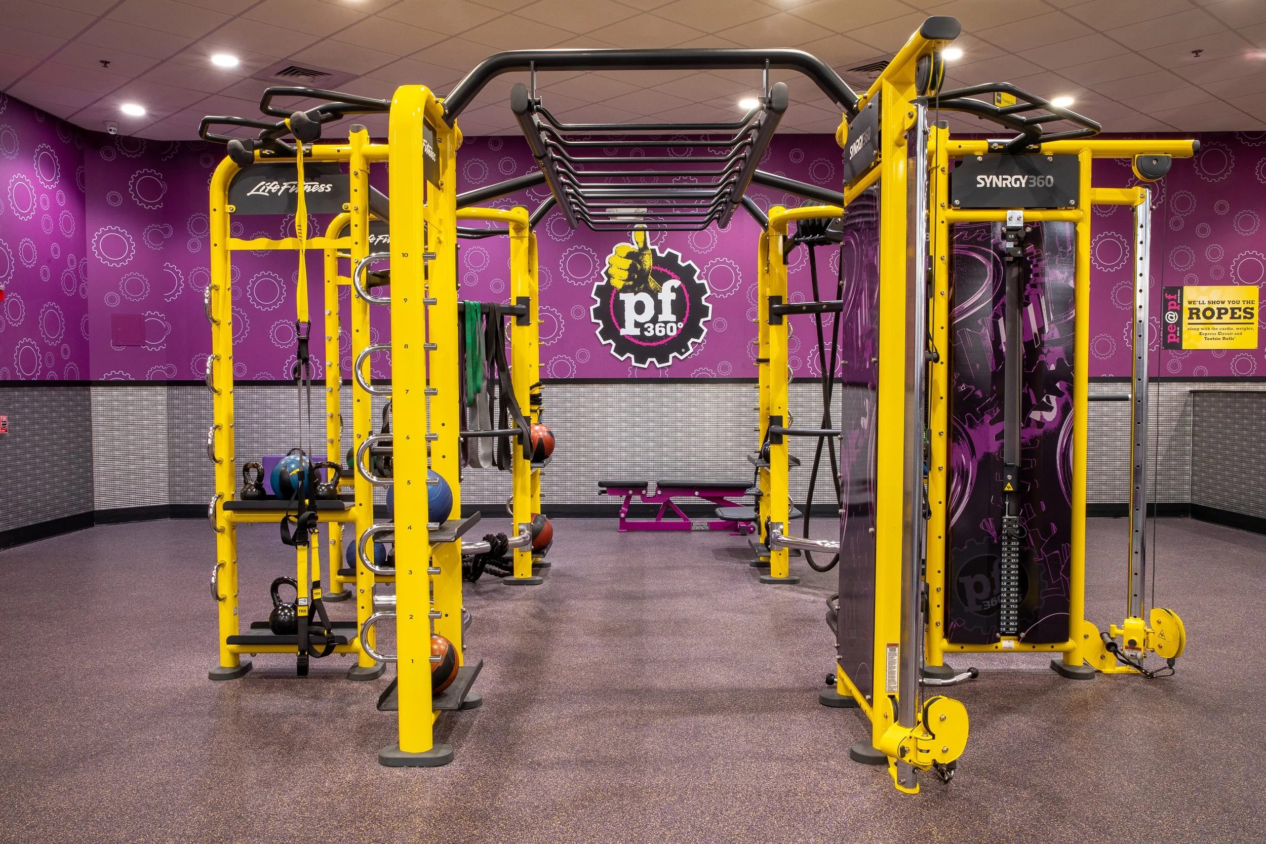 A gym with purple walls decorated with gear illustrations. Yellow and black workout equipment, including a squat rack, kettlebells, and medicine balls, are arranged throughout the room. The background features a large wall graphic with gear illustrat