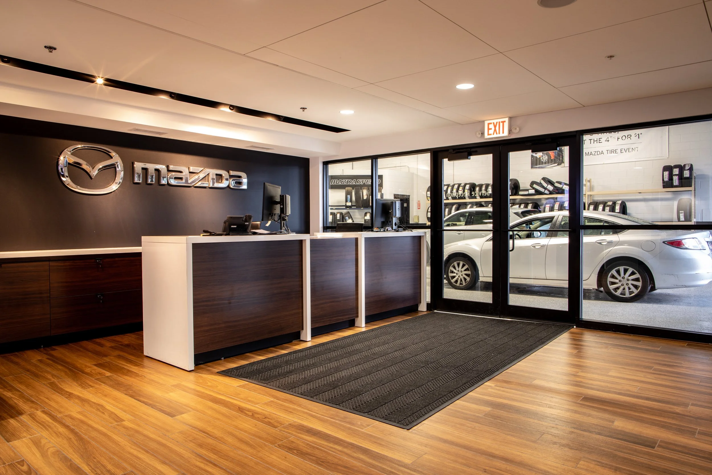 Mazda dealership reception area with a black wall featuring a silver Mazda logo and the word 'Mazda.' There are two computer workstations behind the reception desk, and a glass door reveals a car parked outside along with shelves of tires. An exit si