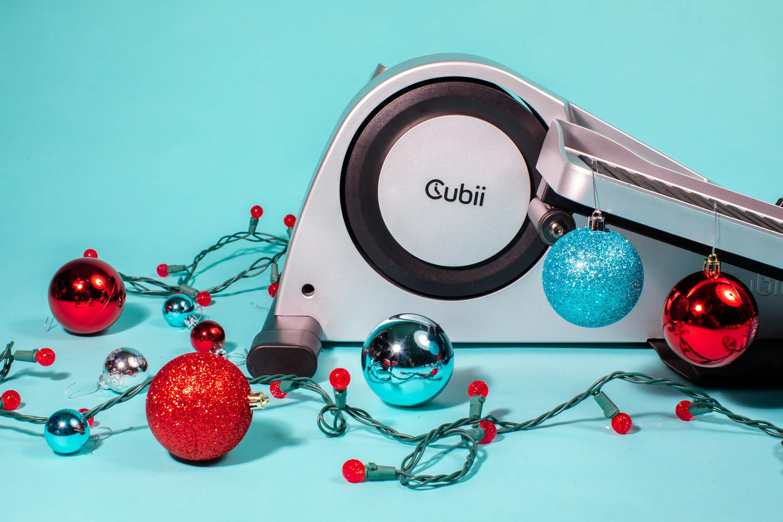A Cubii under-desk elliptical machine surrounded by red, blue, and silver Christmas ornaments and string lights with red bulb covers, on a light blue background.