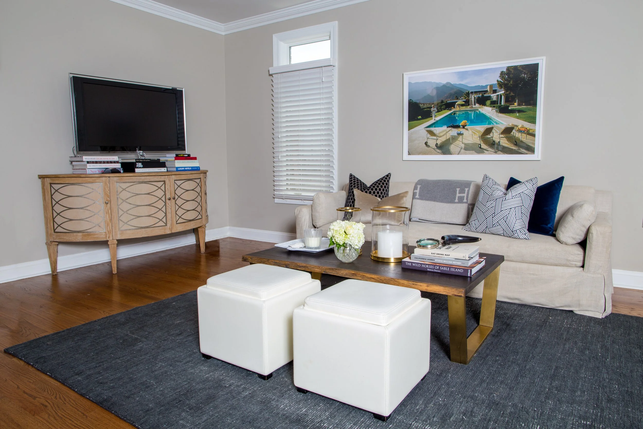 Living room with a beige sofa decorated with patterned and velvet cushions, a wooden coffee table with books and candles, a dark rug, a black flat-screen TV on a wooden stand, a window with blinds, and a large framed photo of an outdoor pool scene on