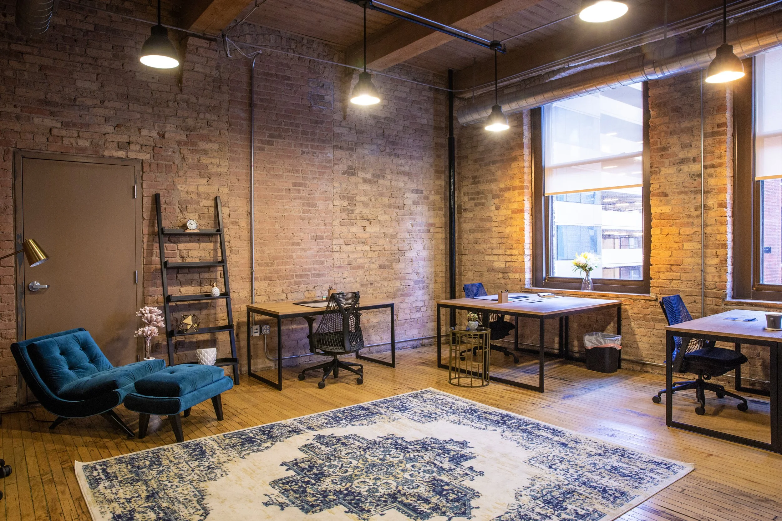 Interior of a modern office space with exposed brick walls, large windows, wooden ceiling, and various tables and chairs. There is a blue tufted sofa and a patterned rug on the wooden floor, with decorative plants and lamps.