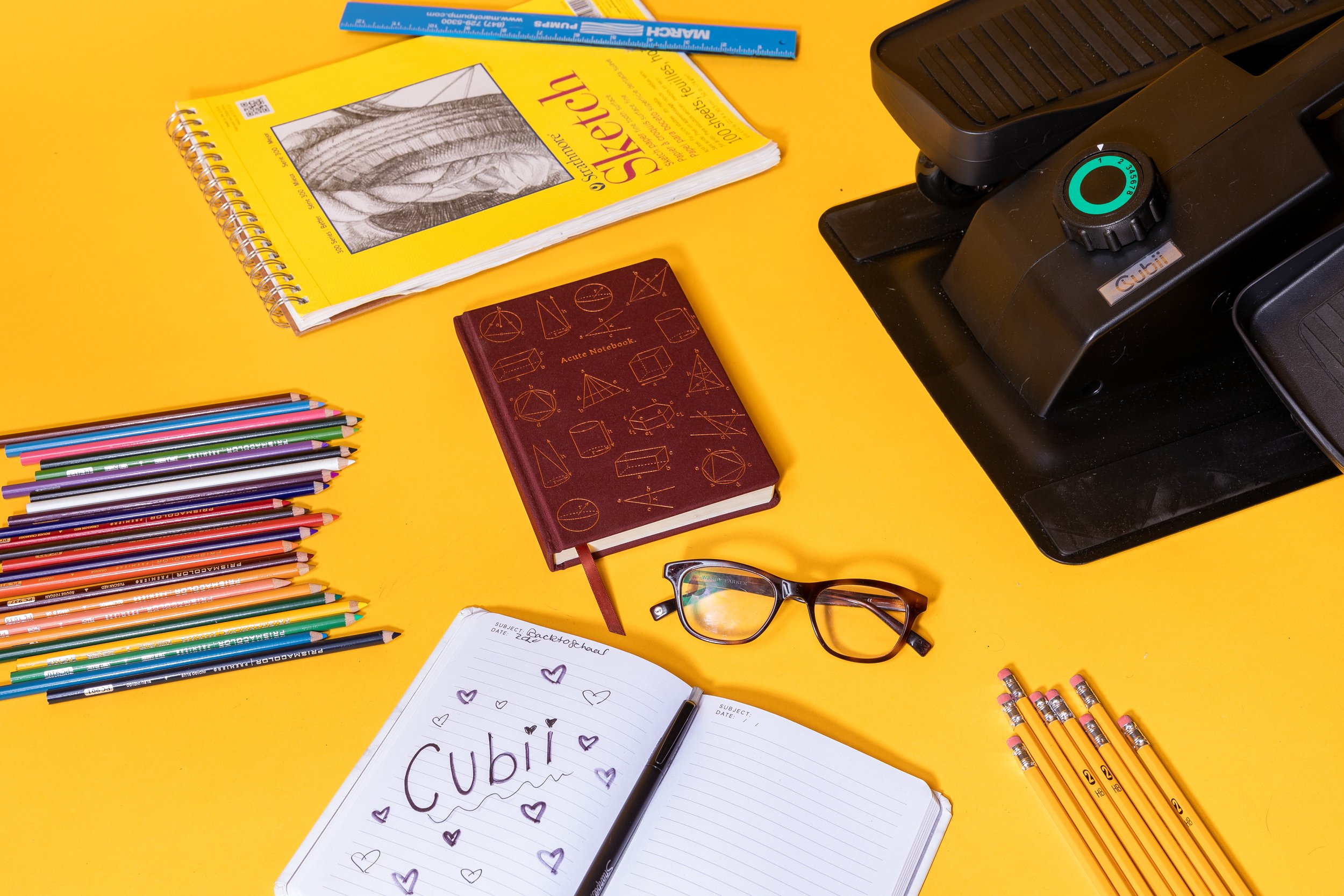 A yellow table surface holds various stationery items, including a stack of colored pencils, a purple notebook with geometric designs, a yellow workbook with a drawing of hands on the cover, a blue ruler, a pair of glasses, a black pen, and a white n