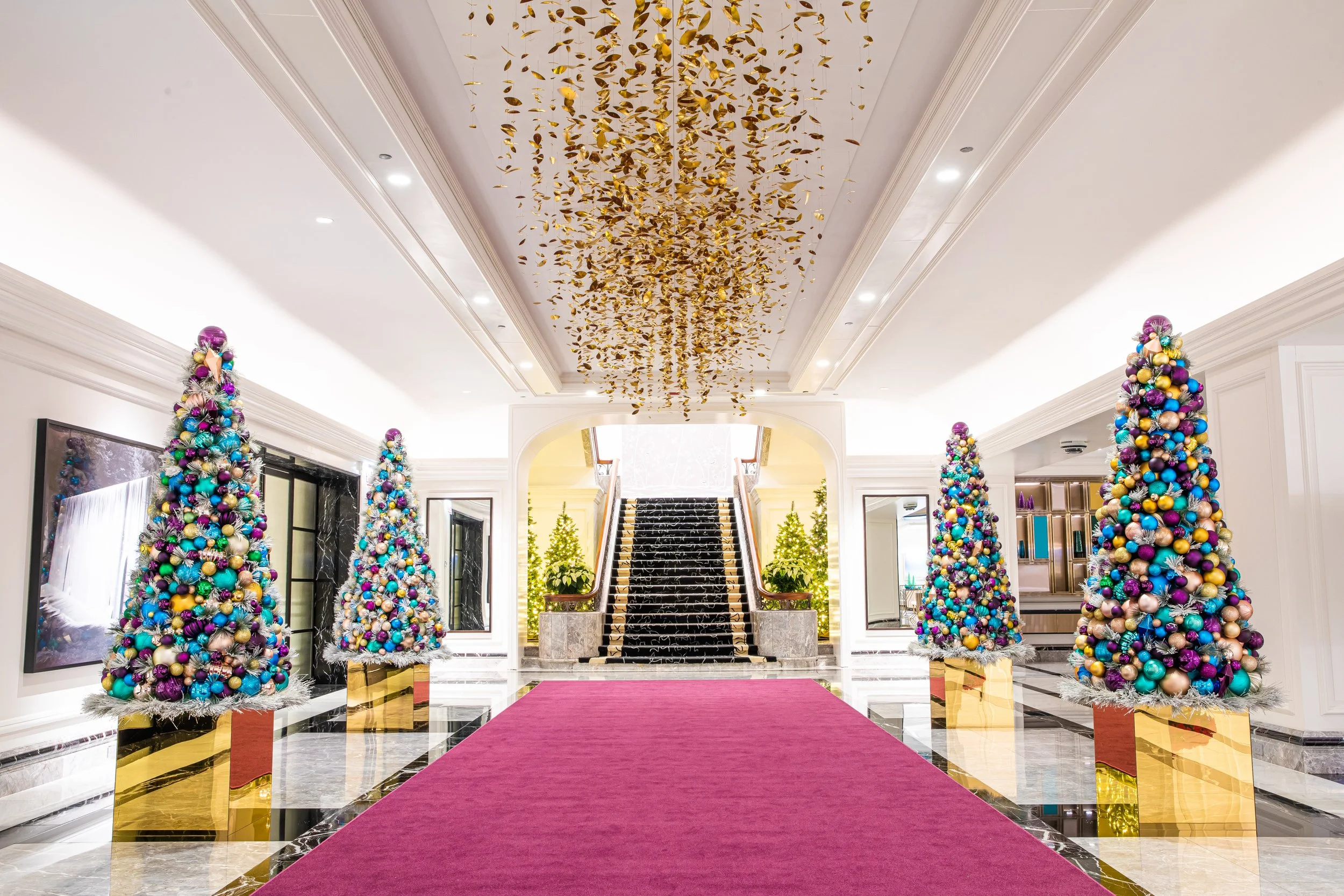 Elegantly decorated hotel lobby with a red carpet leading to a staircase, surrounded by colorful Christmas trees and a gold leaf ceiling decoration.