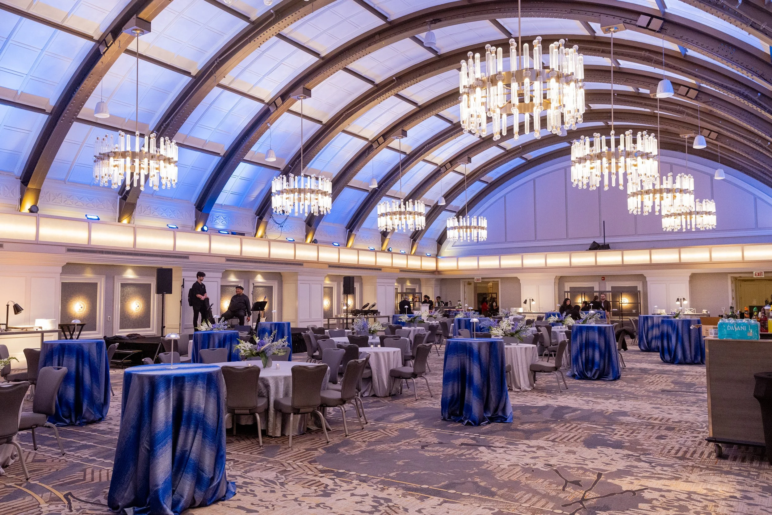 Elegant event space with round tables covered in blue tablecloths, white floral centerpieces, and modern chandeliers hanging from the arched ceiling, with some people preparing for an event.